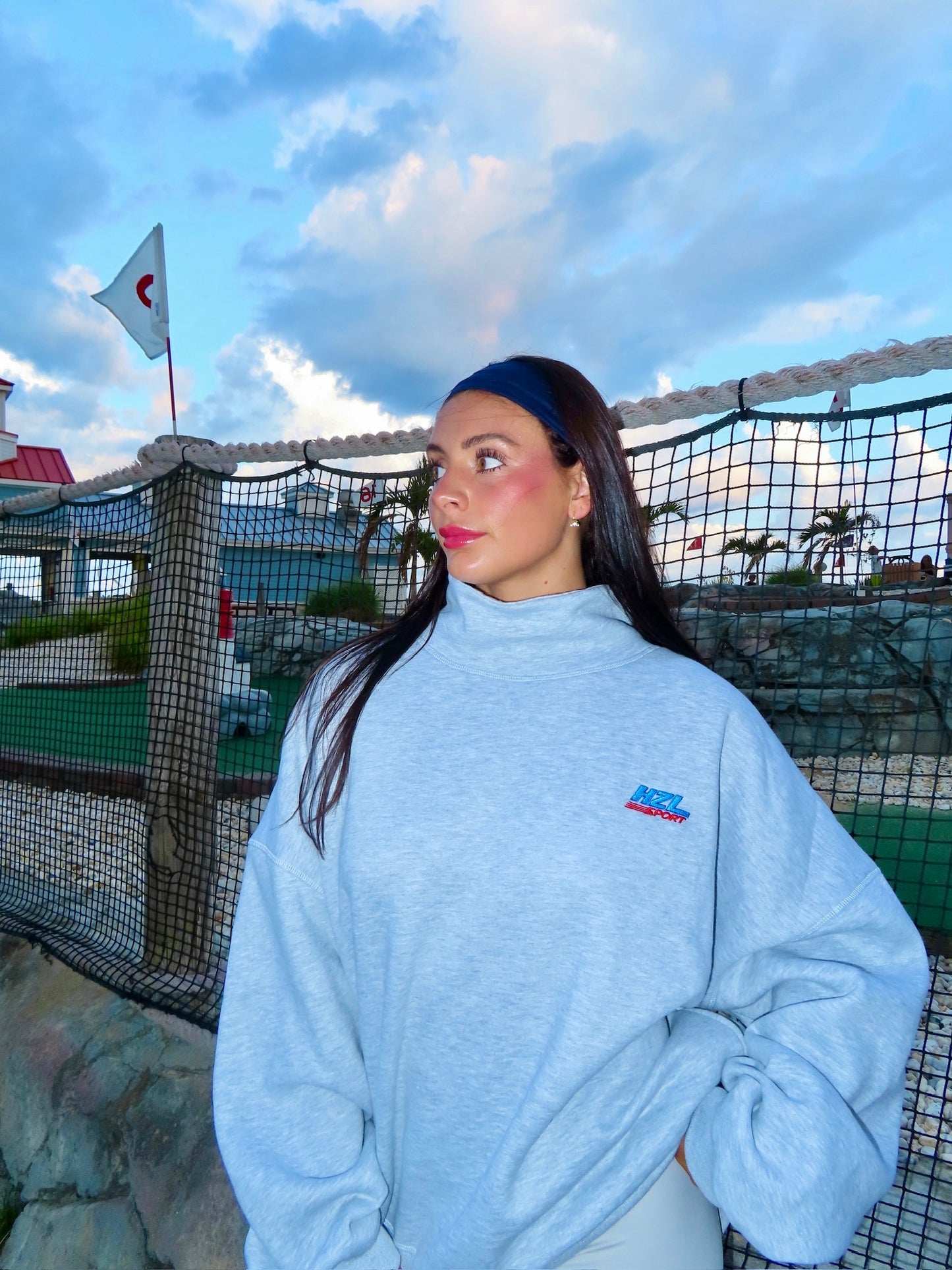 woman wearing a grey mockneck sweatshirt with a small logo standing outdoors near a netted fence at dusk