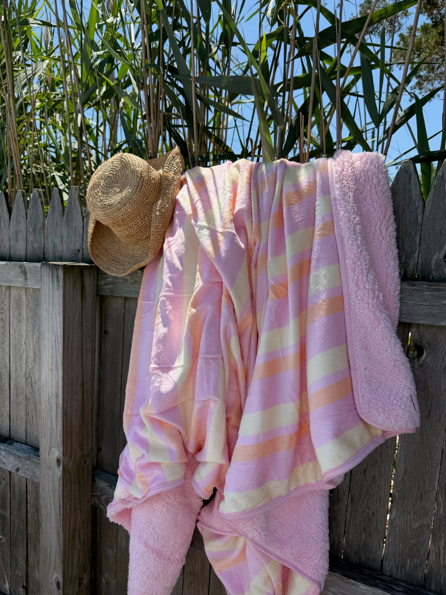 Soft pastel striped blanket draped over a wooden fence with a straw sun hat hanging beside it under a sunny sky