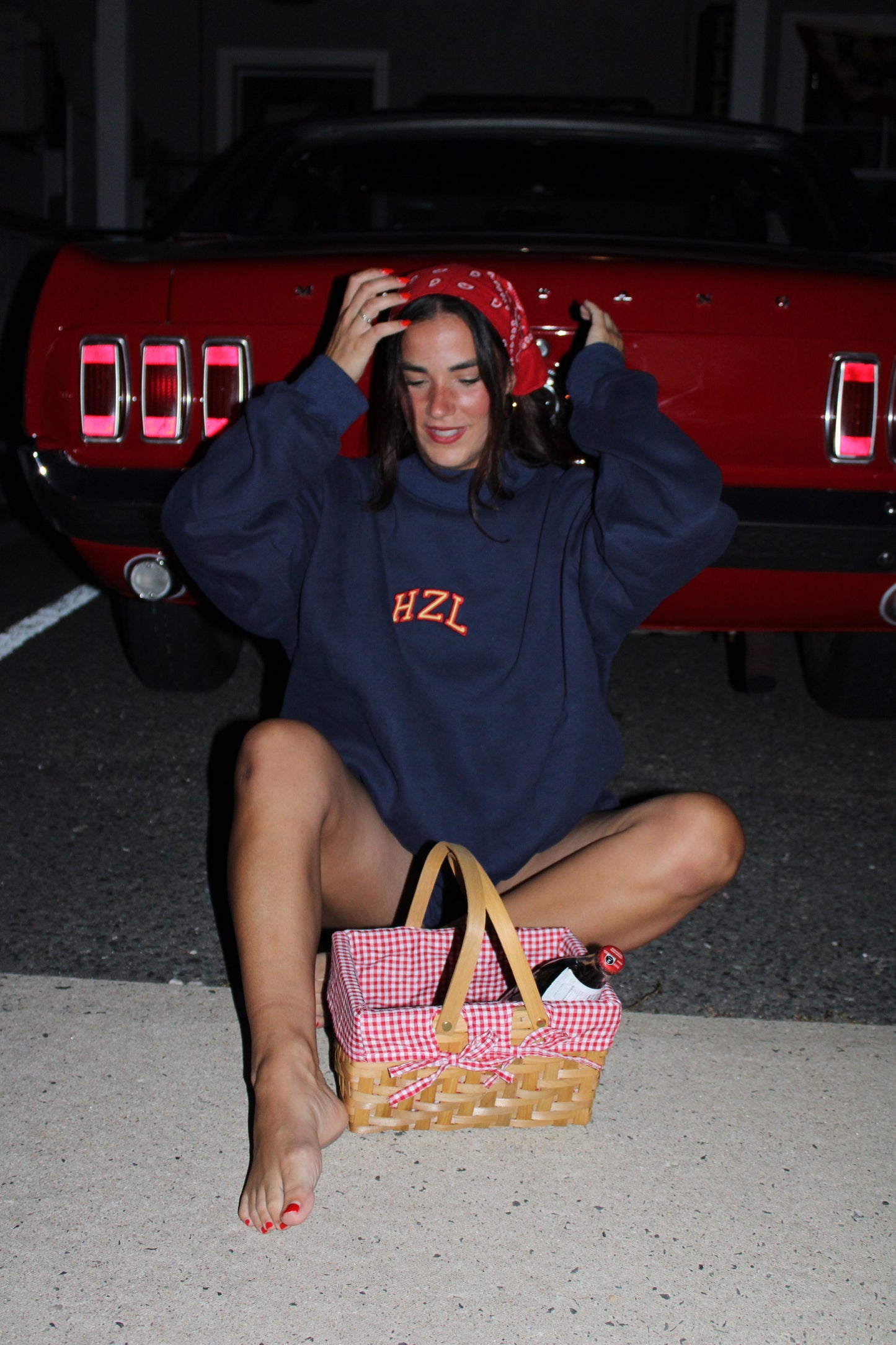 Woman sitting barefoot on pavement at night wearing a navy HZL sweatshirt and red bandana, with a picnic basket in front of her and a red vintage car behind.