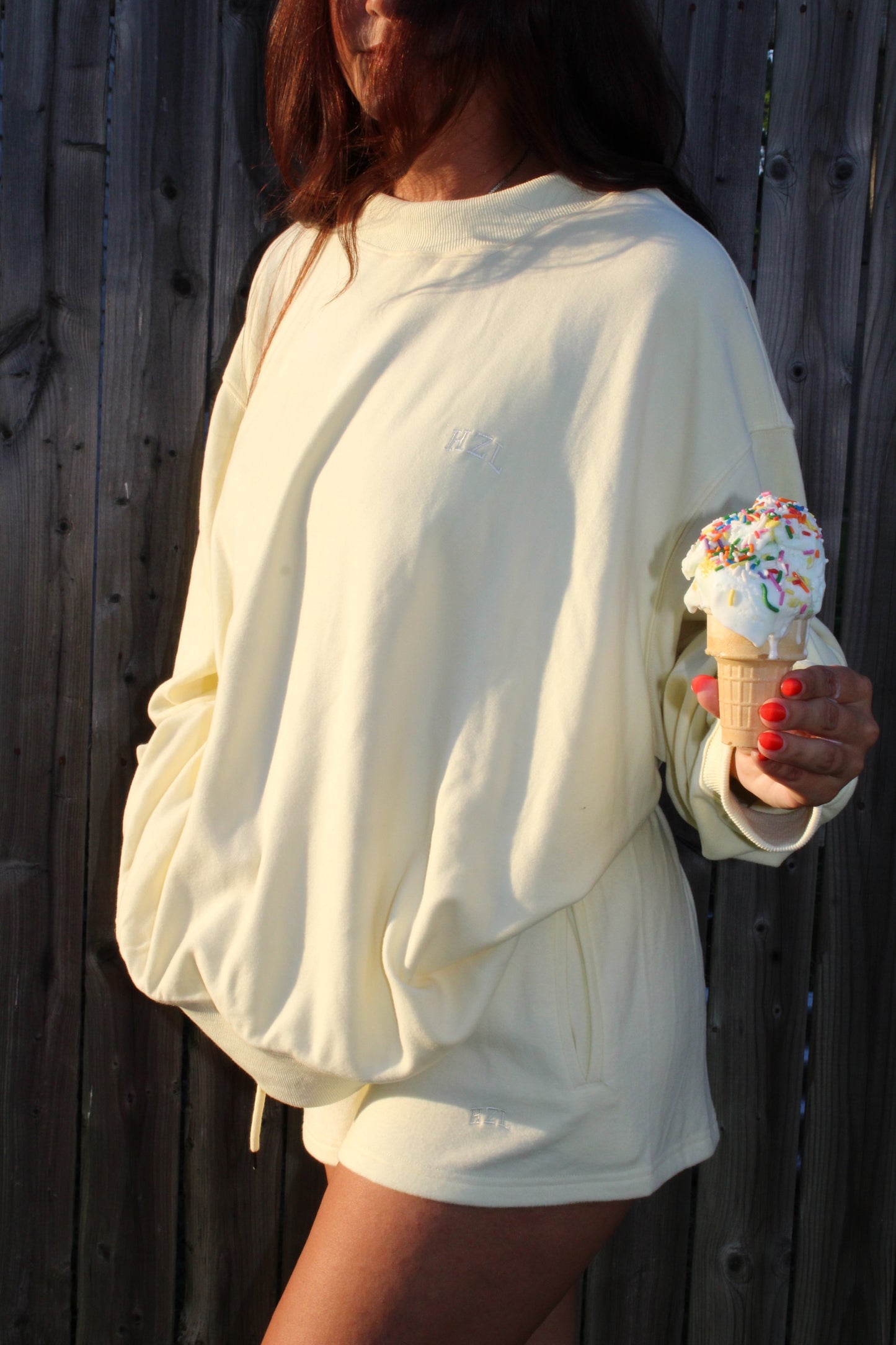 Woman wearing a soft yellow oversized mockneck and shorts set holding an ice cream cone with sprinkles against a wooden fence background