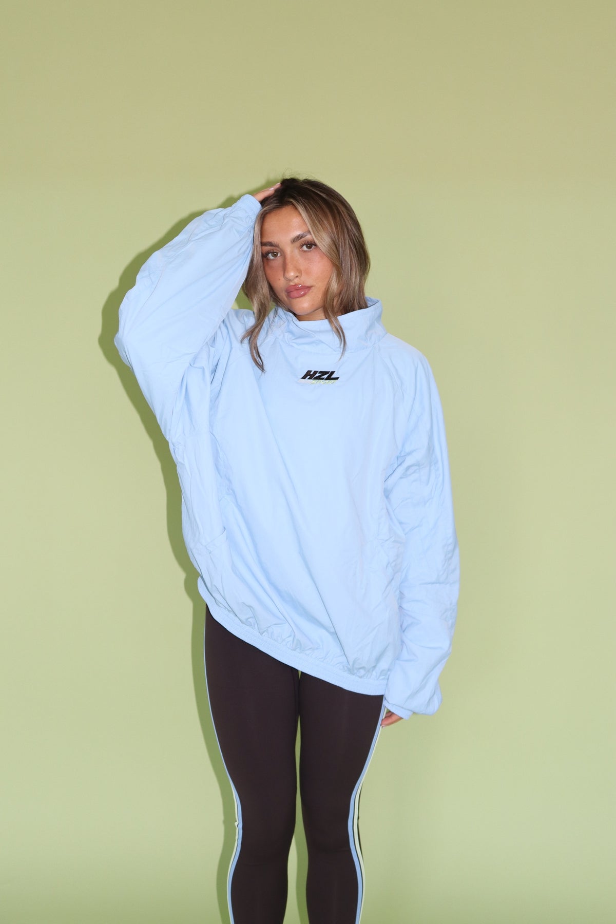 Model wearing a light blue HZL windbreaker with black leggings against a green background