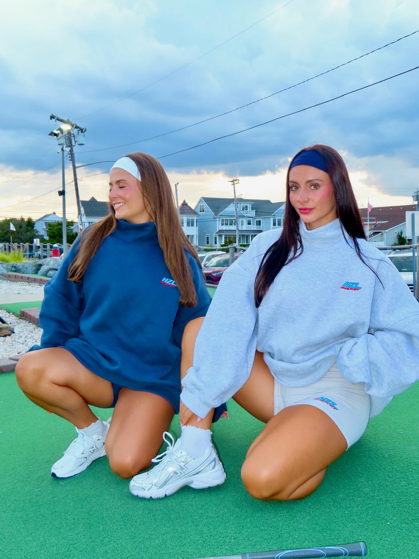 Two women wearing sporty oversized sweatshirts and fitted shorts, sitting outdoors on green turf near a mini golf course under a cloudy sky.