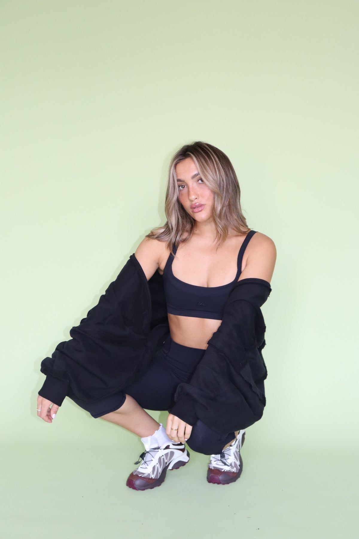 Woman crouching in a black warm up shrug with an athletic top and sneakers