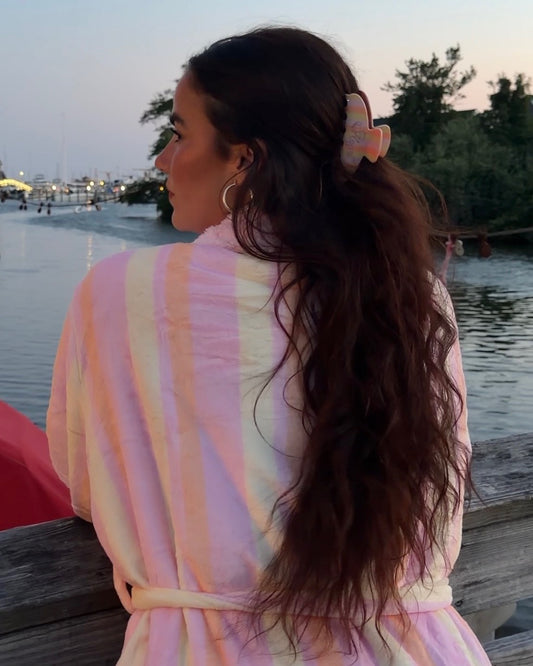 Woman with long wavy hair wearing a pastel striped robe and a colorful hair clip by the water at sunset