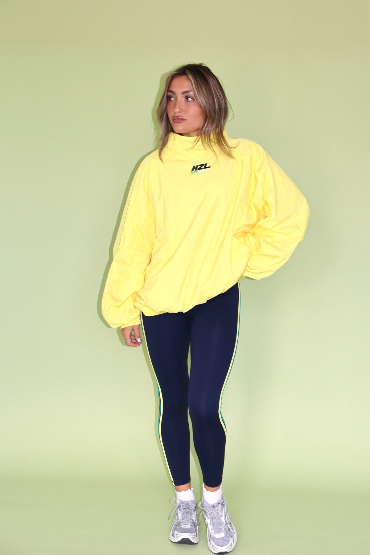 Model wearing a yellow HZL Sport windbreaker with navy leggings and sneakers against a green background