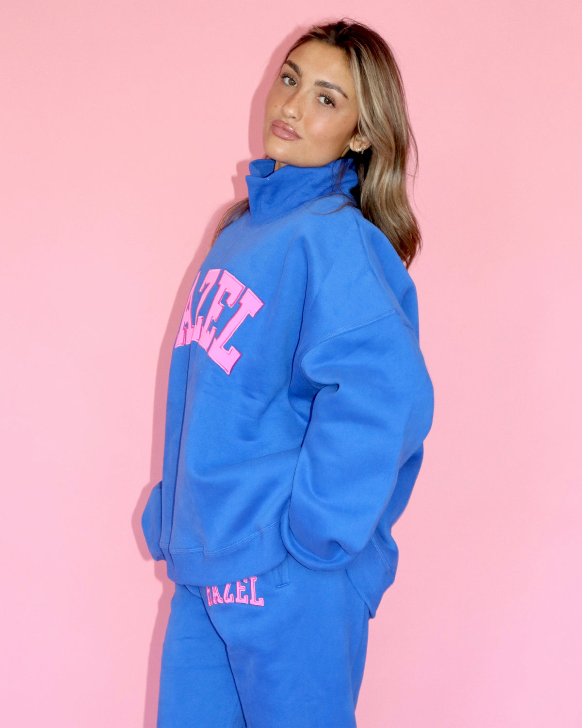 Model wearing a bright blue high-neck sweatshirt with pink lettering against a pink background