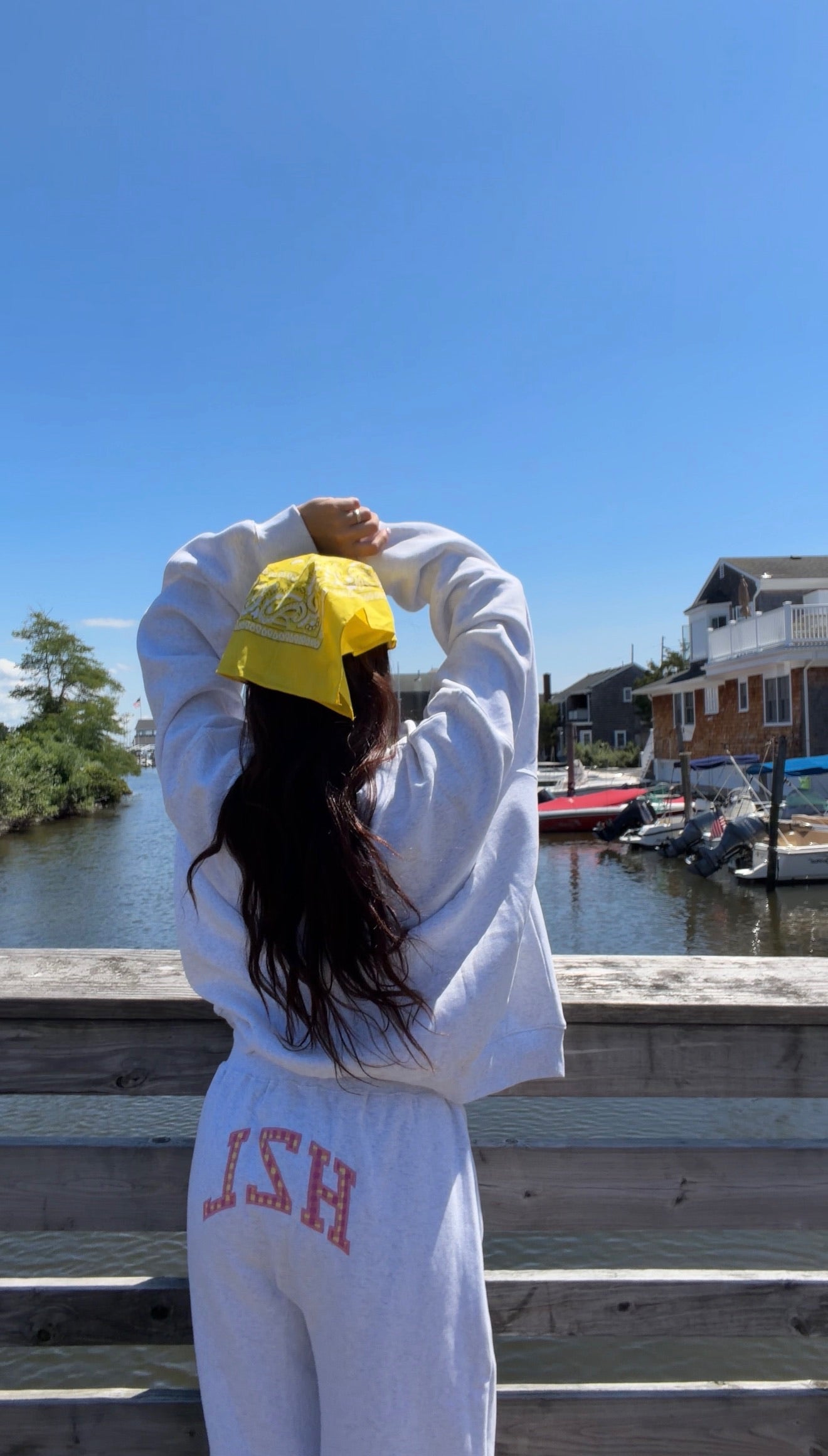Woman with long dark hair wearing light gray casual pants with a printed logo and a yellow bandana, standing by a waterfront railing under a clear blue sky.