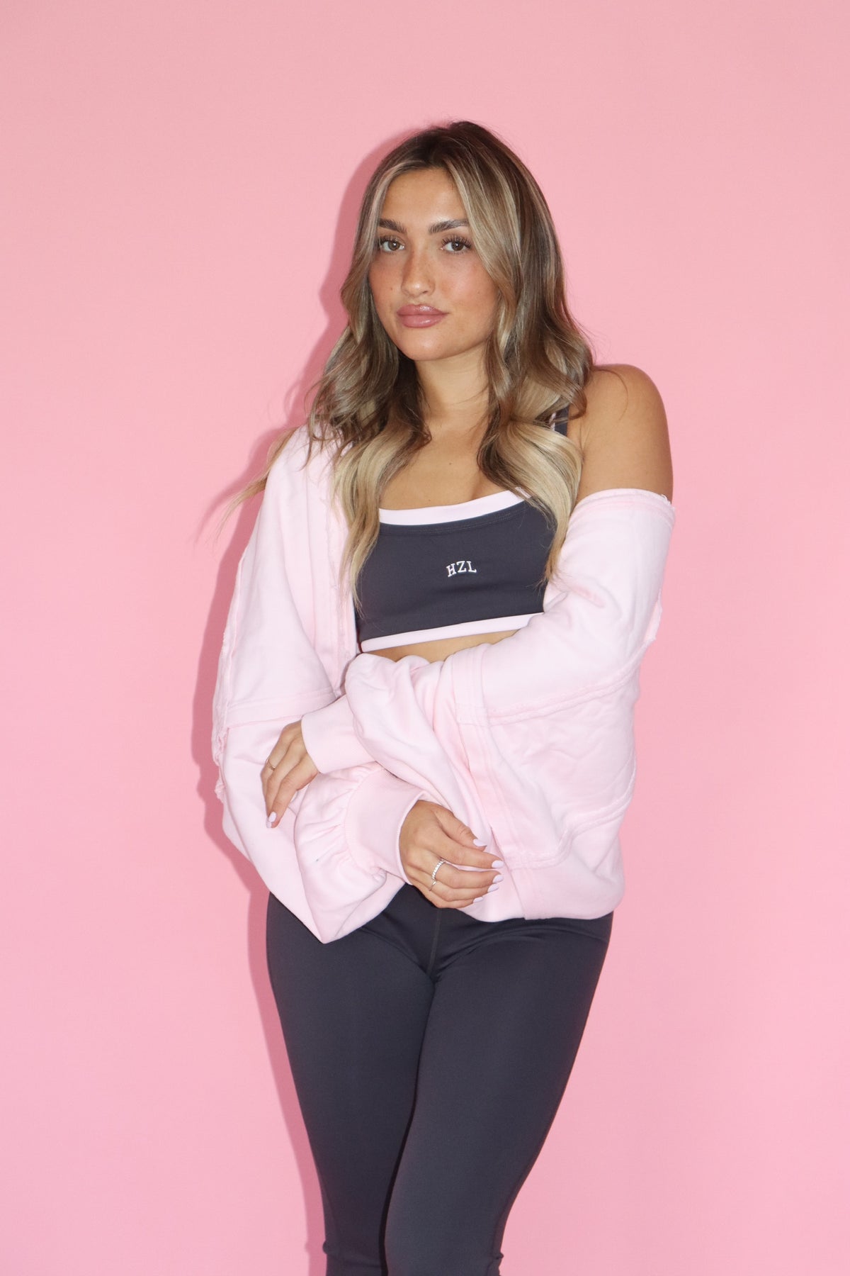 Woman wearing a pink warm up shrug over a black crop top and leggings against a pink background