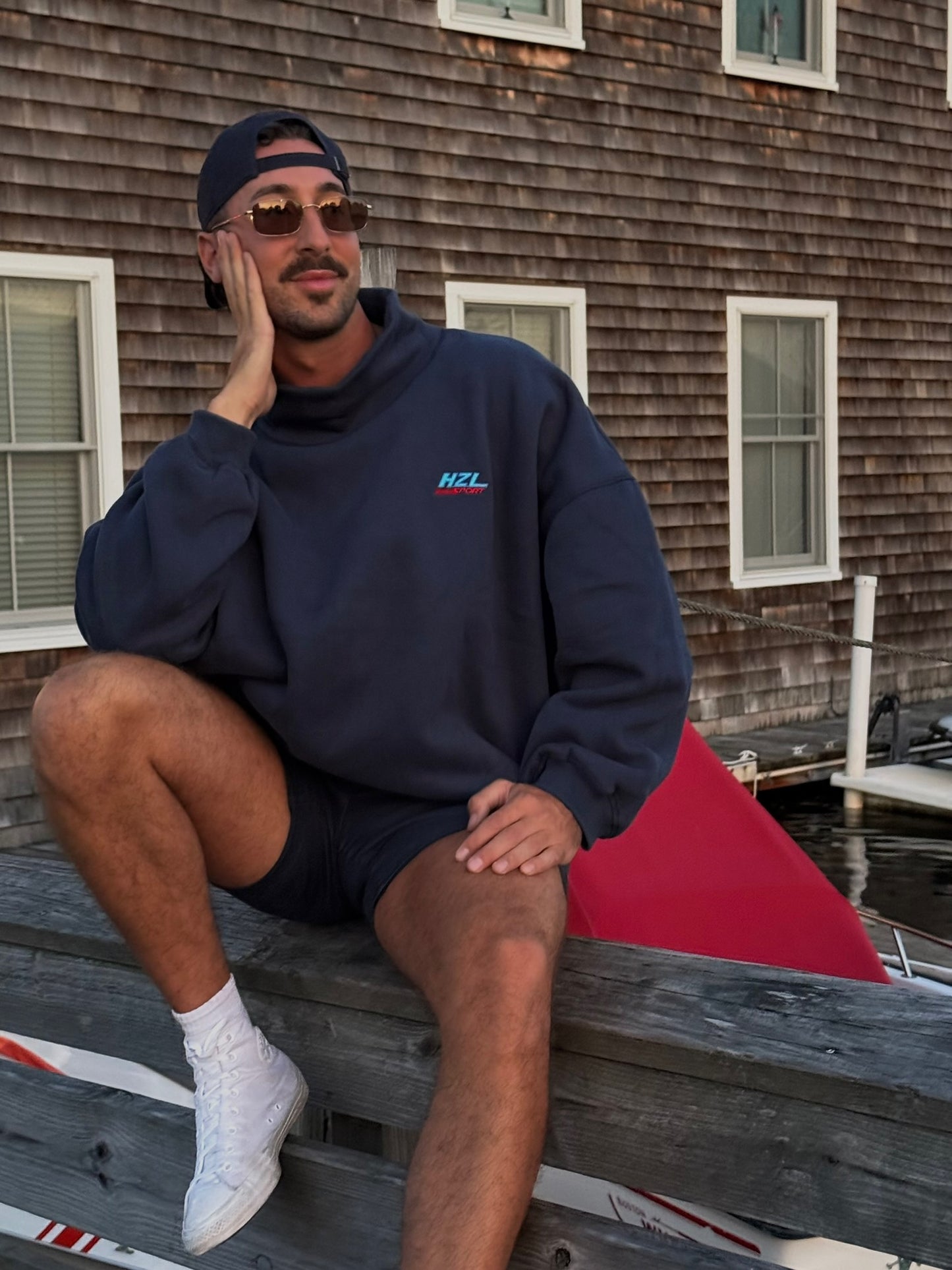 man wearing navy mockneck sweatshirt sitting casually on wooden dock near water with sunglasses and cap