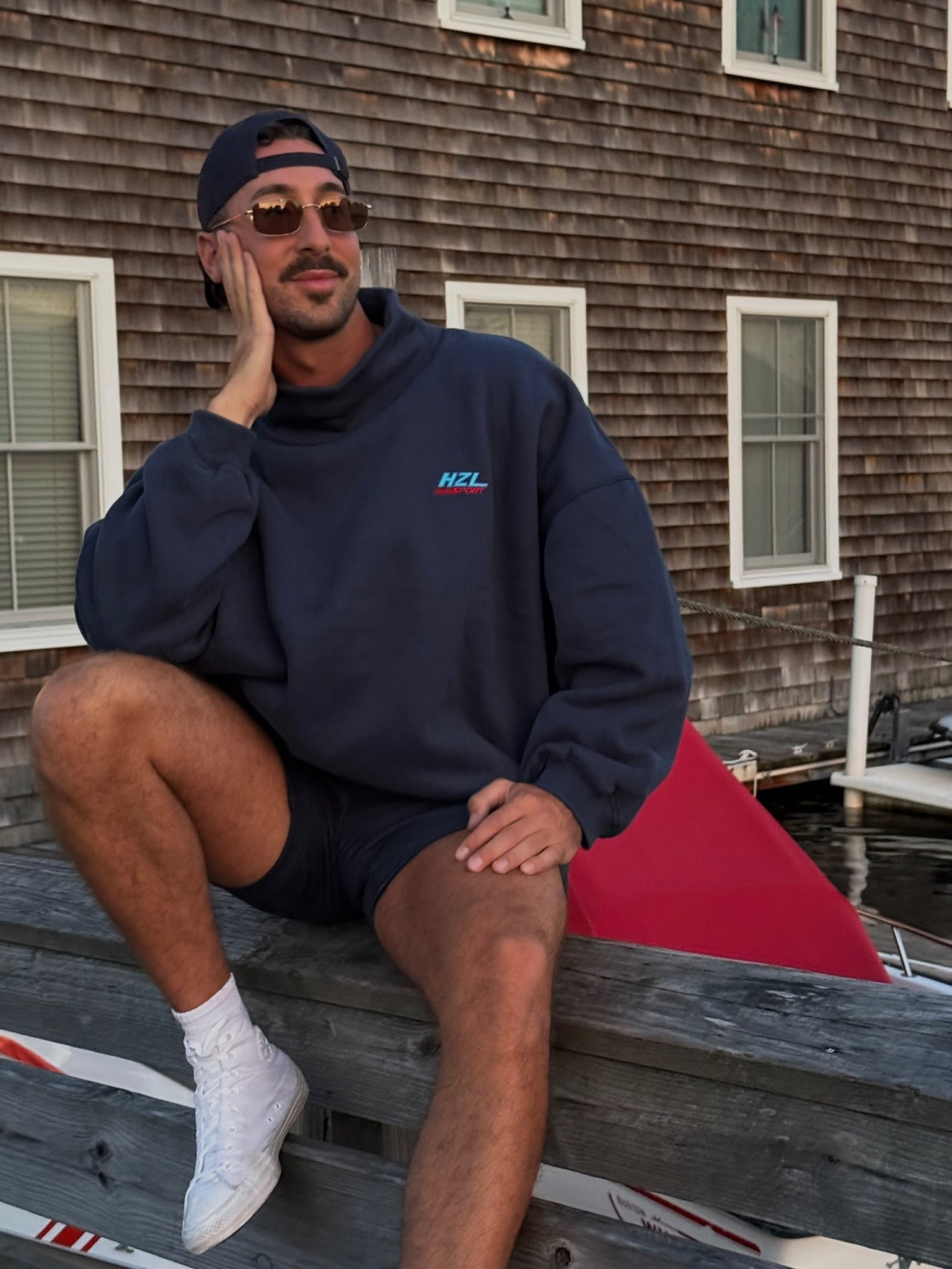 man wearing navy mockneck sweatshirt sitting casually on wooden dock near water with sunglasses and cap