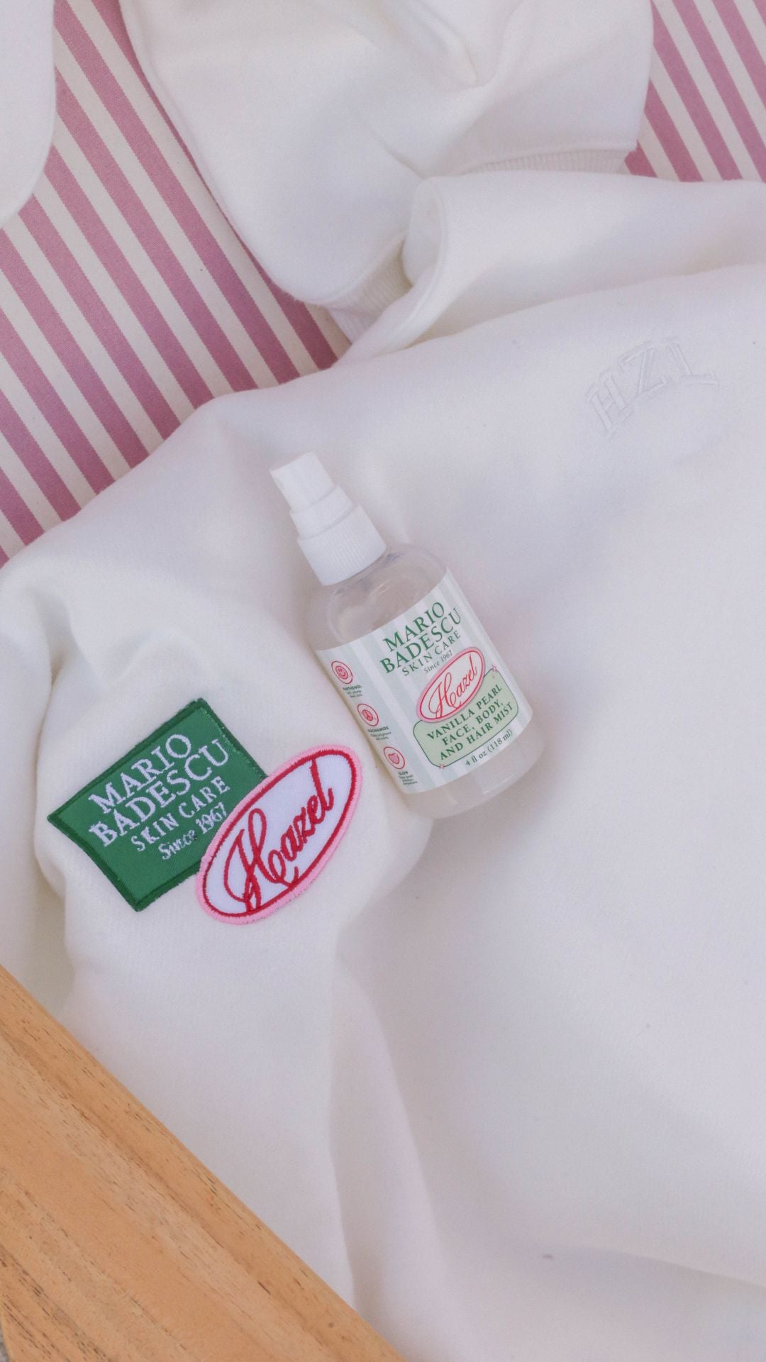 Mario Badescu Vanilla Pearl and Herb Mist bottle on a white embroidered sweatshirt with pink striped background