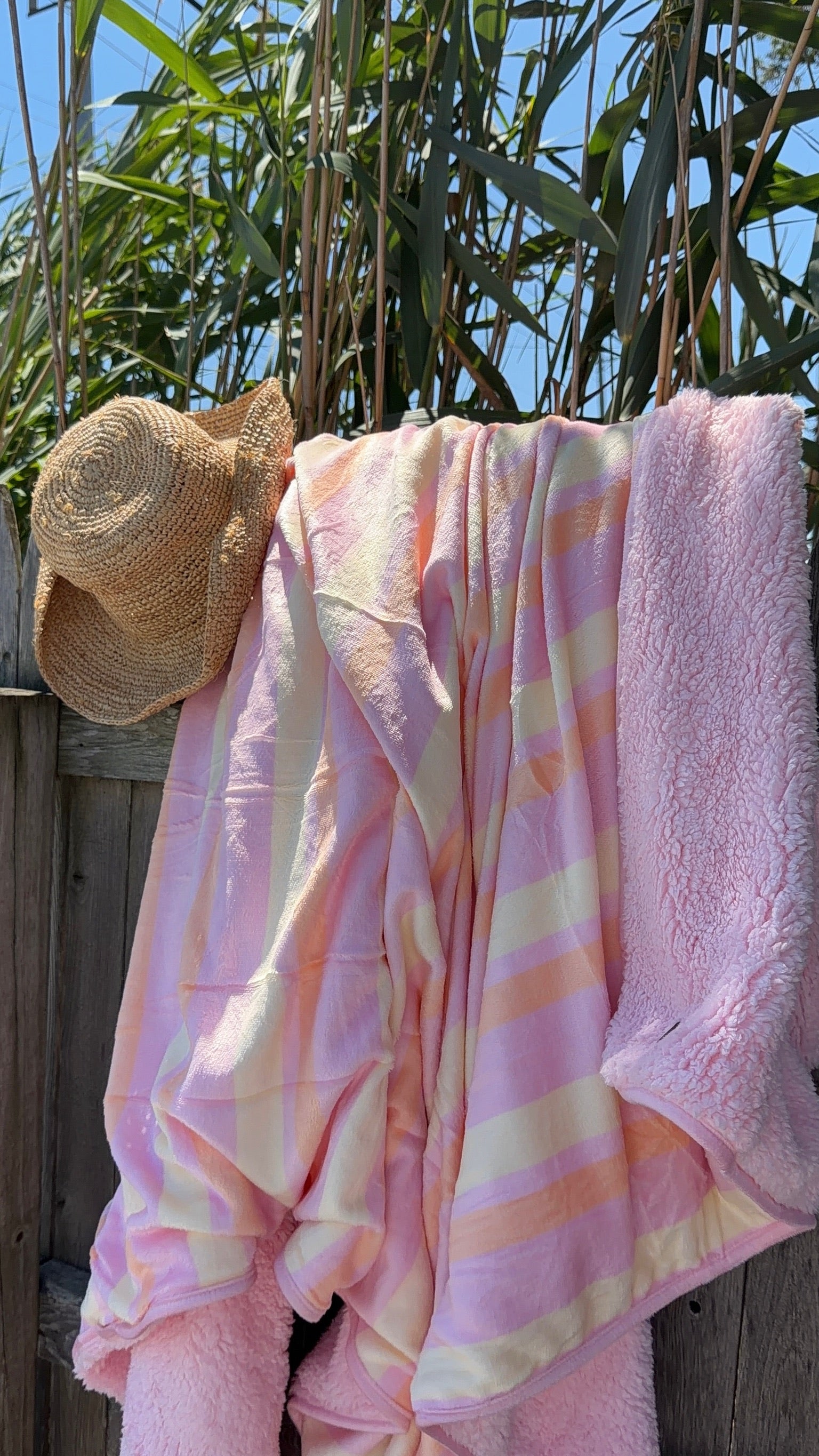 Soft pastel pink and cream striped blanket draped over a wooden fence with a straw hat hanging beside it, set against green foliage and blue sky.