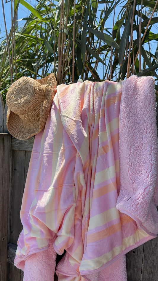 Soft pastel pink and cream striped blanket draped over a wooden fence with a straw hat hanging beside it, set against green foliage and blue sky.