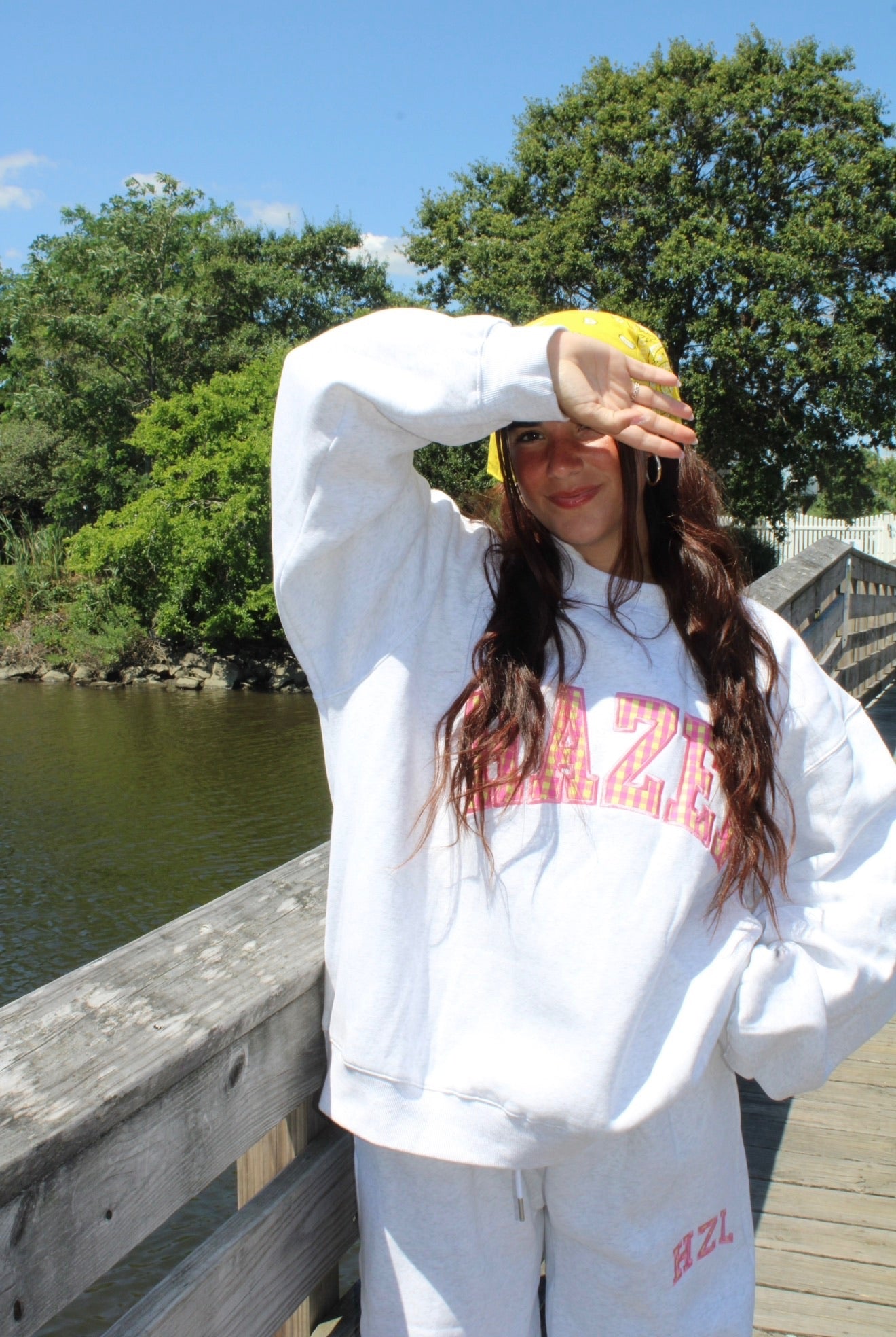 Young woman in casual white sweatshirt and sweatpants on a sunny wooden bridge by the water, shielding her face from sunlight