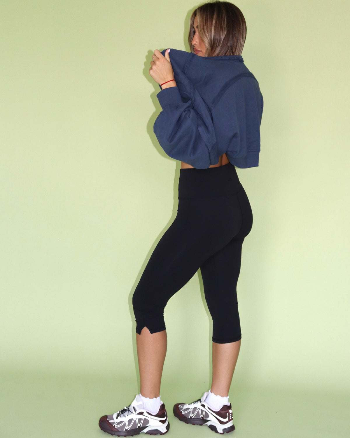 Model wearing a navy cropped warm up shrug with black leggings and sneakers from the back view