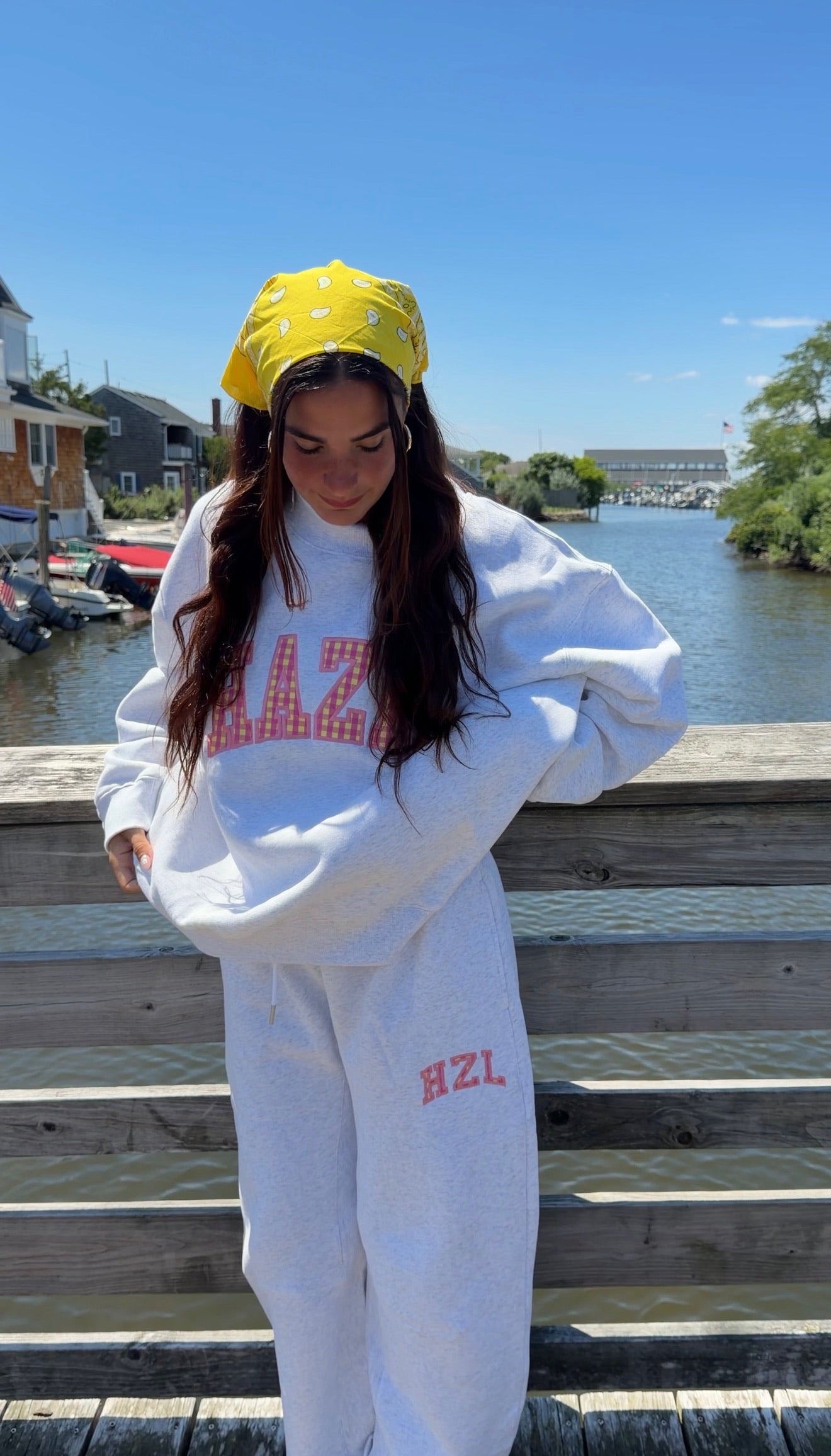 Woman wearing light gray casual outfit with pink lettering, standing on wooden dock by a calm waterway under clear blue sky