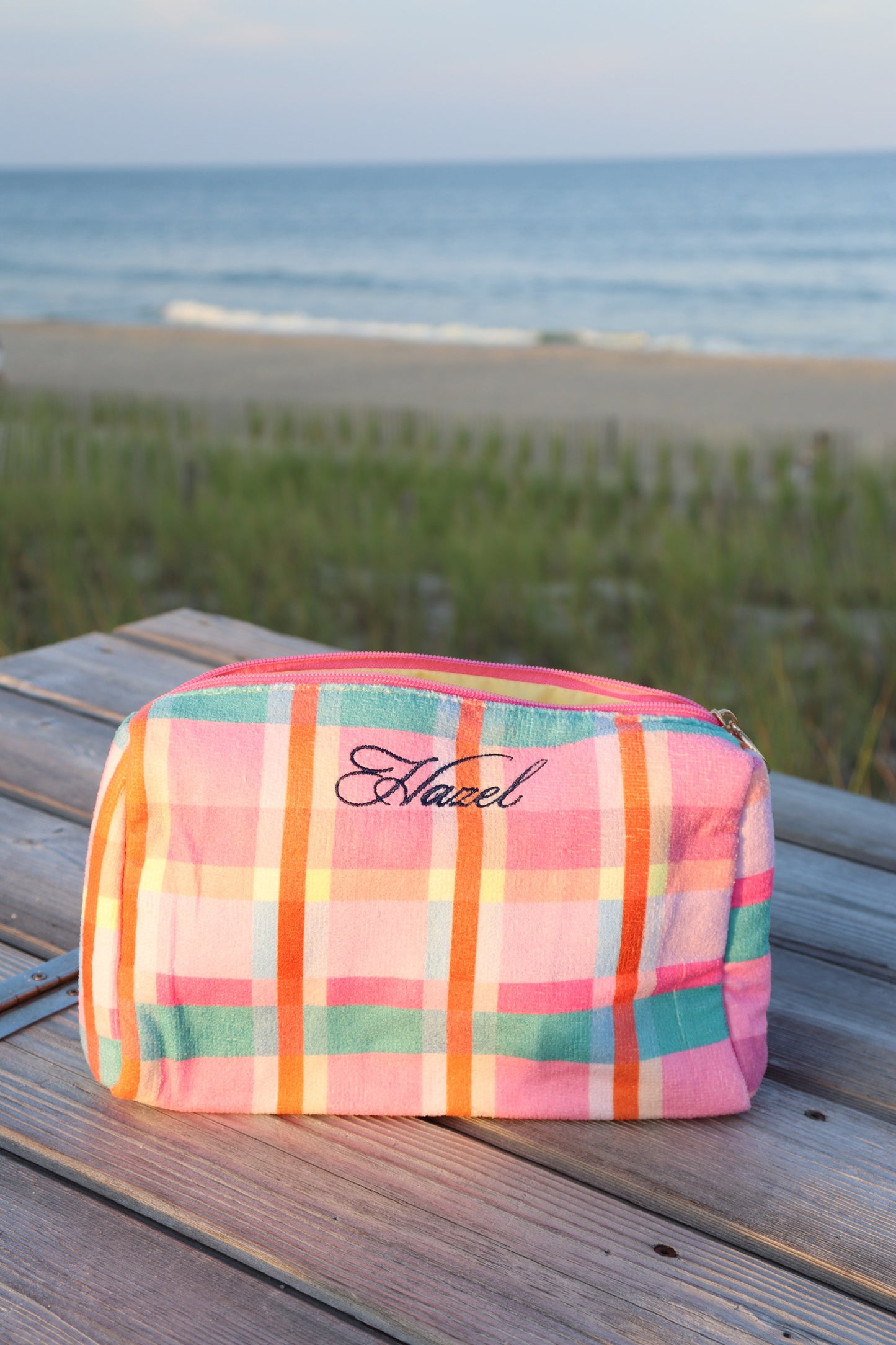 Colorful plaid makeup pouch with Hazel script resting on a wooden beachside boardwalk bench