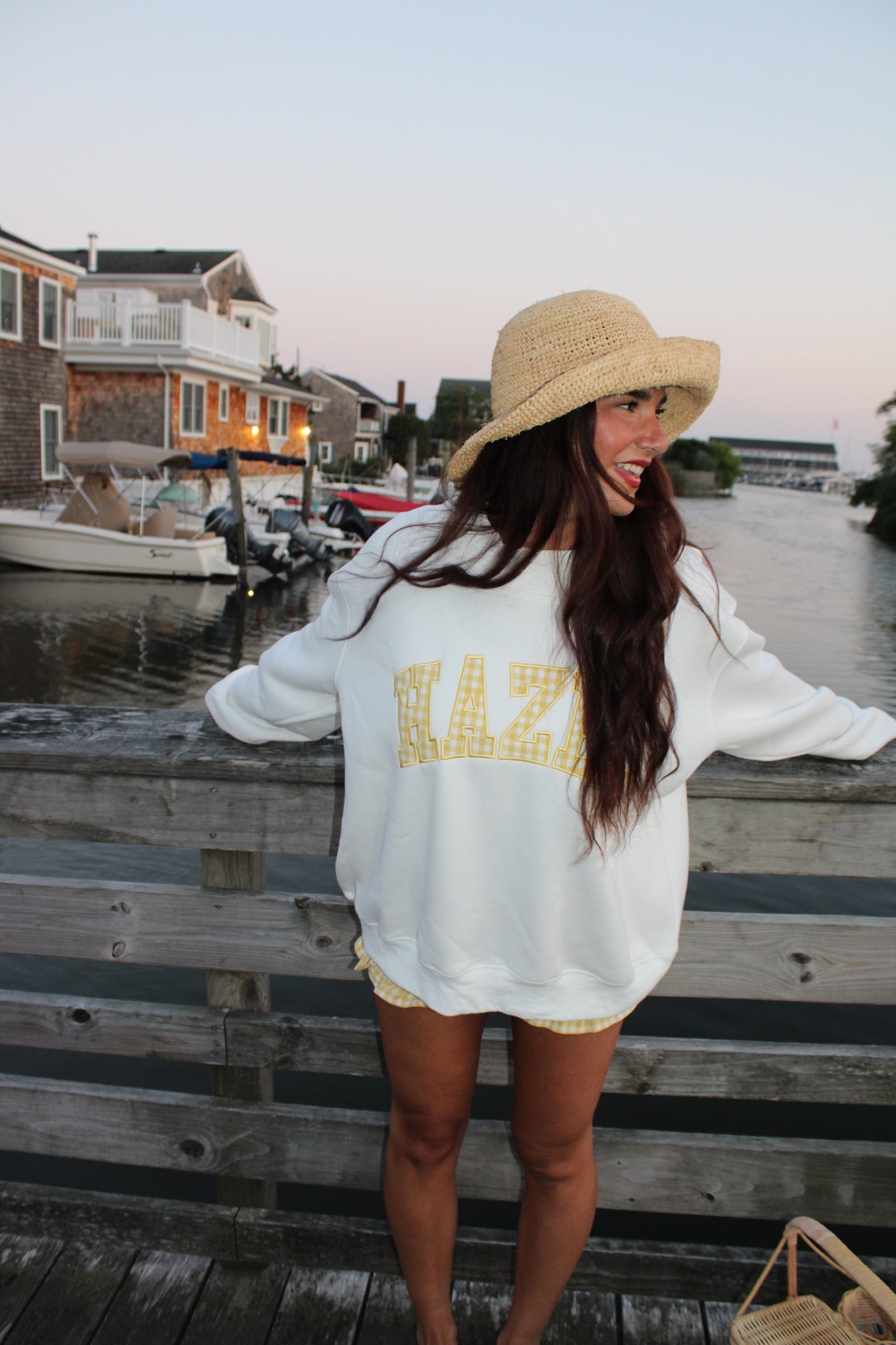 Woman wearing a white sweatshirt with yellow lettering and a straw hat, standing on a wooden dock by the water at sunset.