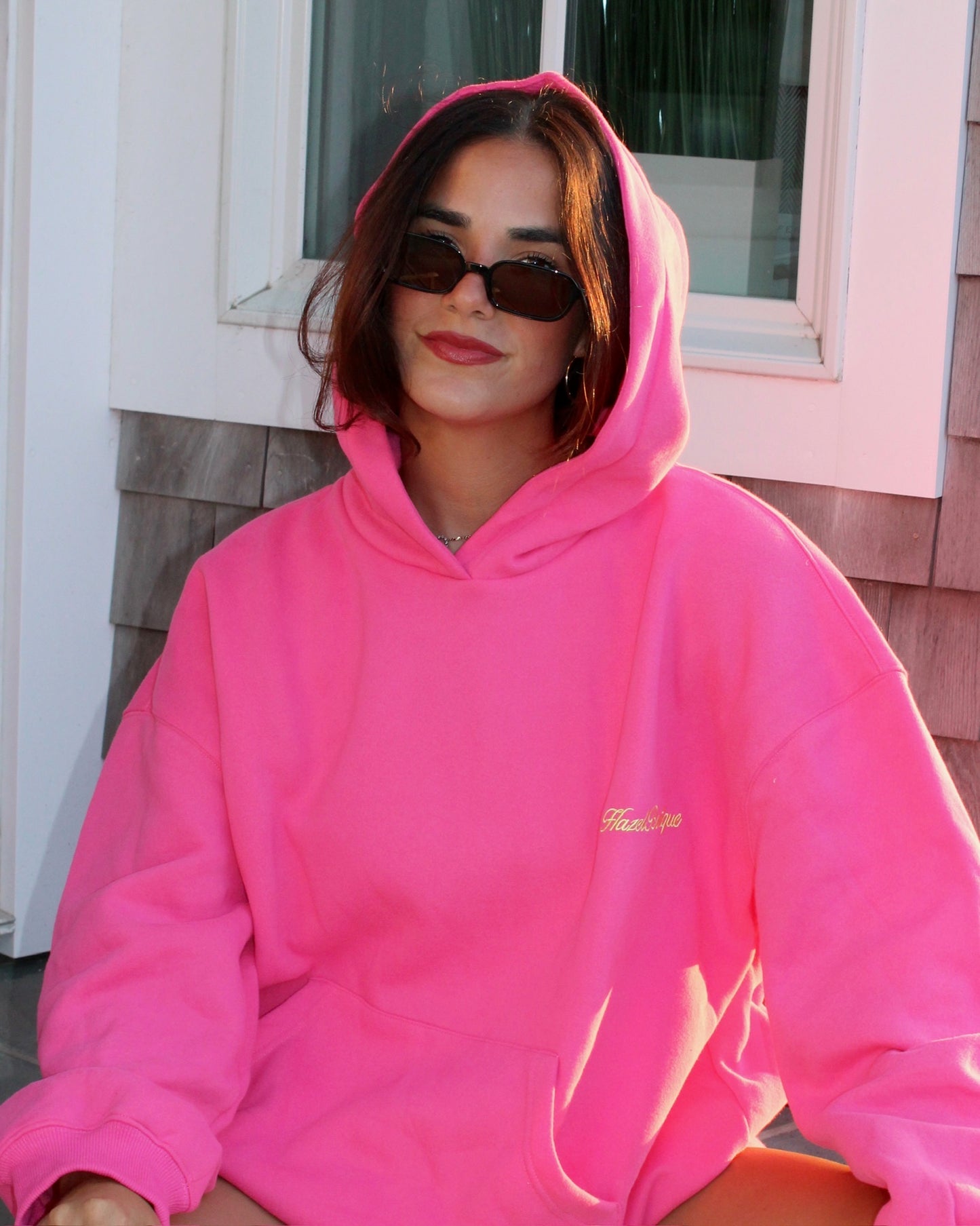 Young woman wearing a bright pink hoodie with small yellow embroidery, paired with dark sunglasses in casual outdoor setting.