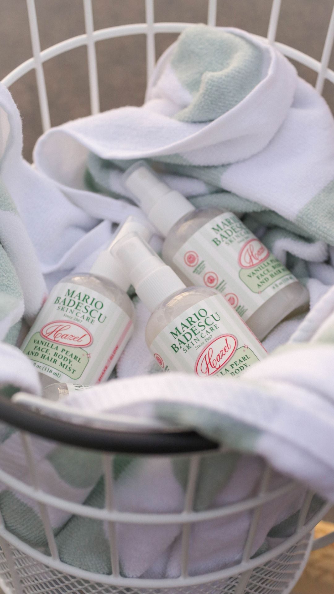 Three bottles of Mario Badescu Vanilla Pearl face, body, and hair mist resting on a white and green towel inside a wire basket.