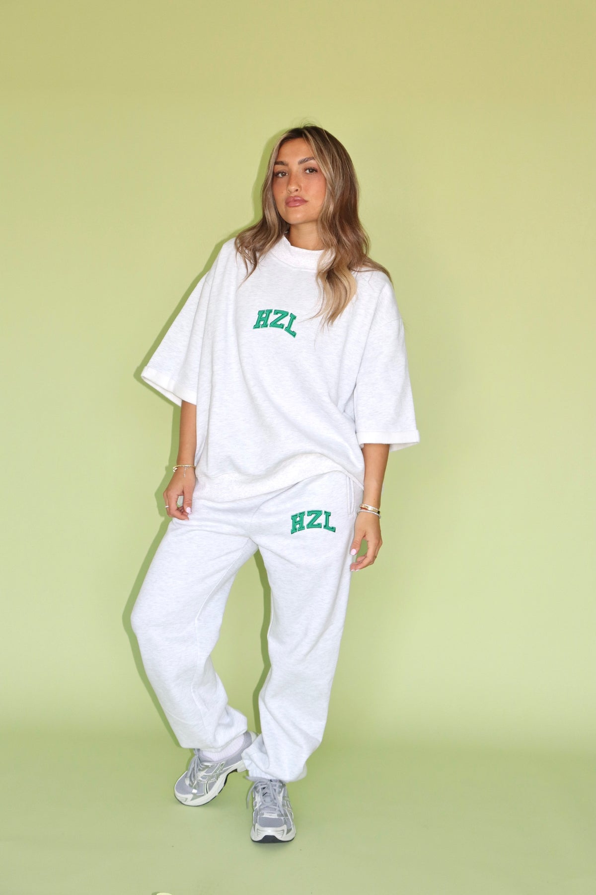 Model wearing white HZL joggers and an oversized tee with green logo against a pastel green backdrop