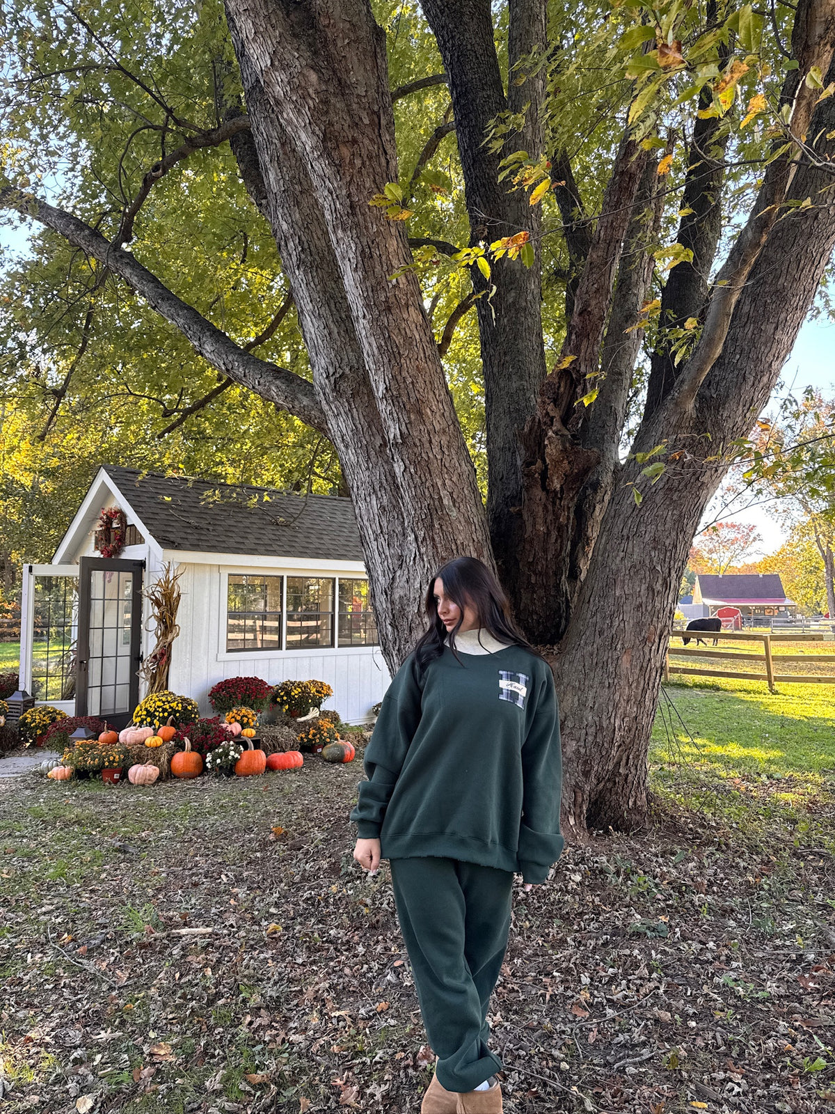 Model wearing a green jogger set outdoors beside a large tree and garden cottage