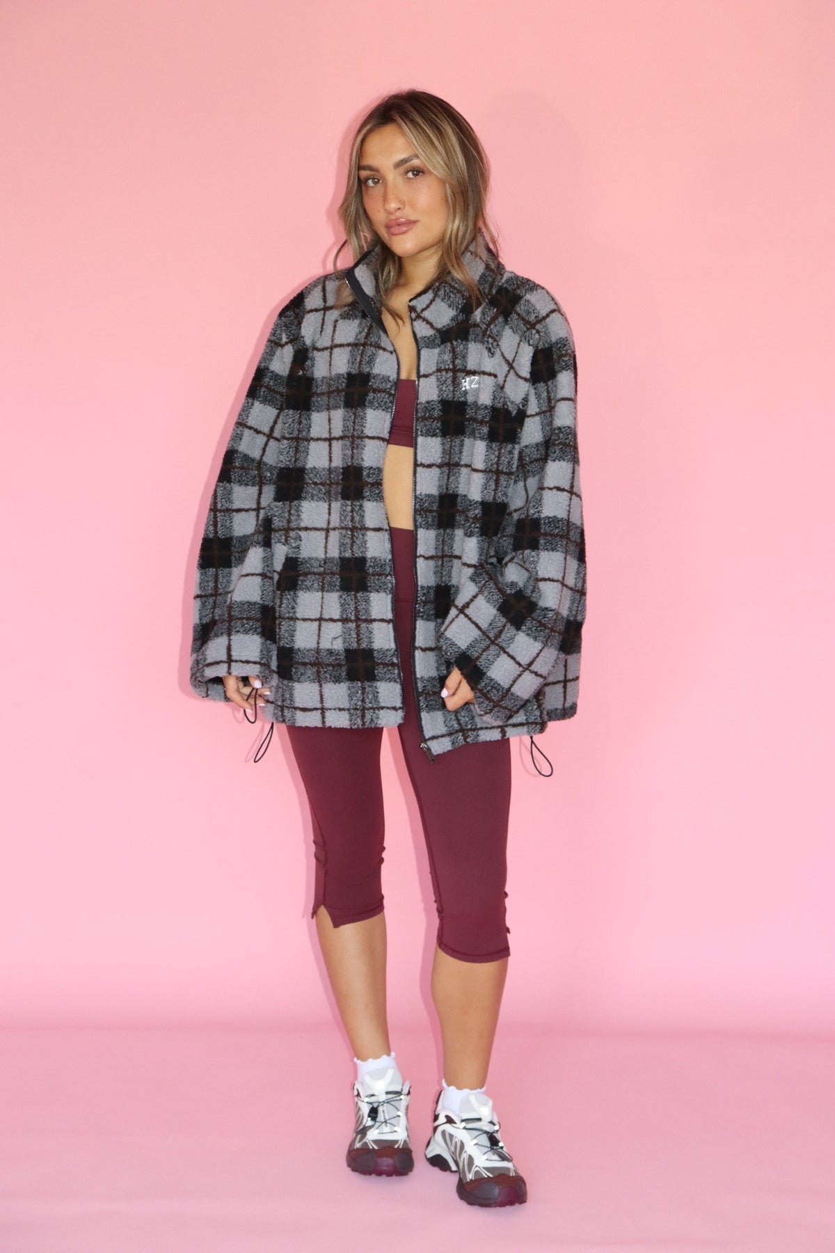 Model wearing a gray and black plaid fleece jacket with burgundy cropped leggings and sneakers on a pink backdrop