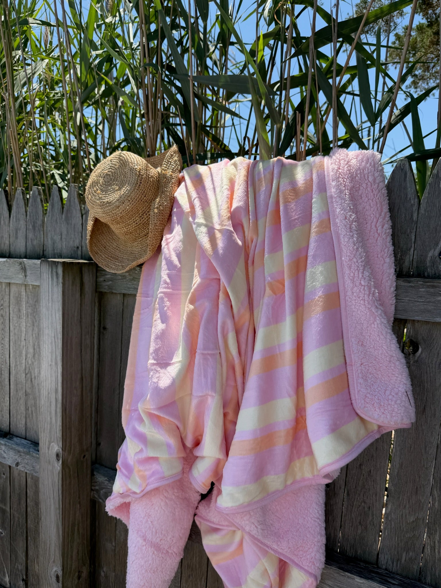 Soft pastel striped blanket draped over a wooden fence with a straw sun hat hanging beside it under a sunny sky