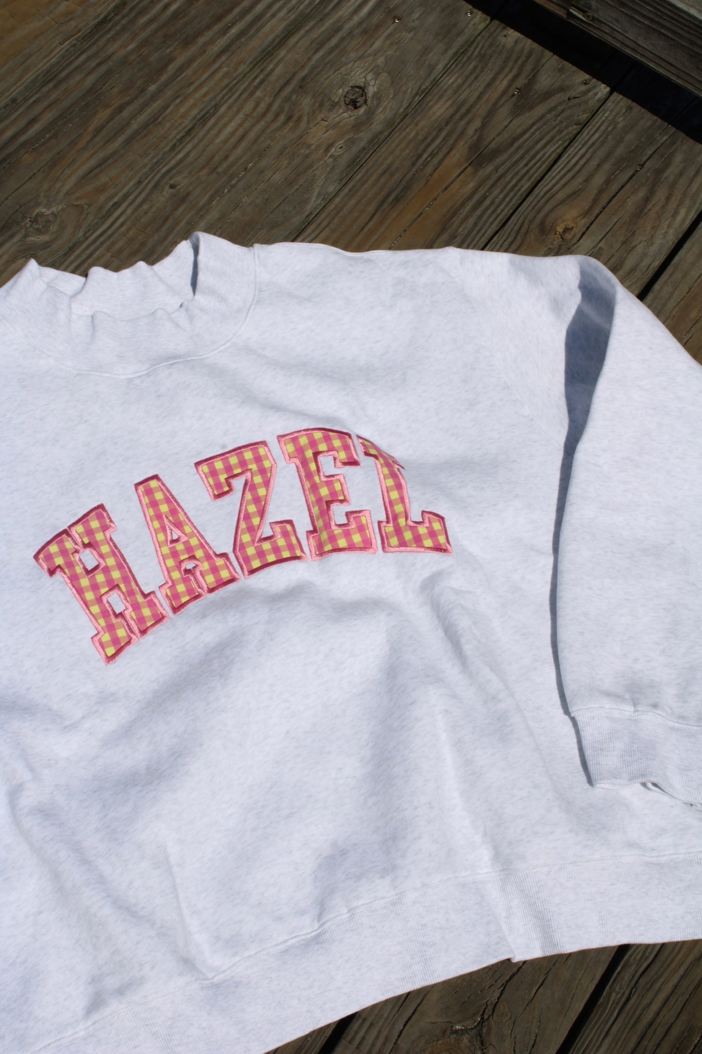 Light gray sweatshirt with pink and yellow gingham patterned letters spelling "HAZEL" laid flat on wooden surface.