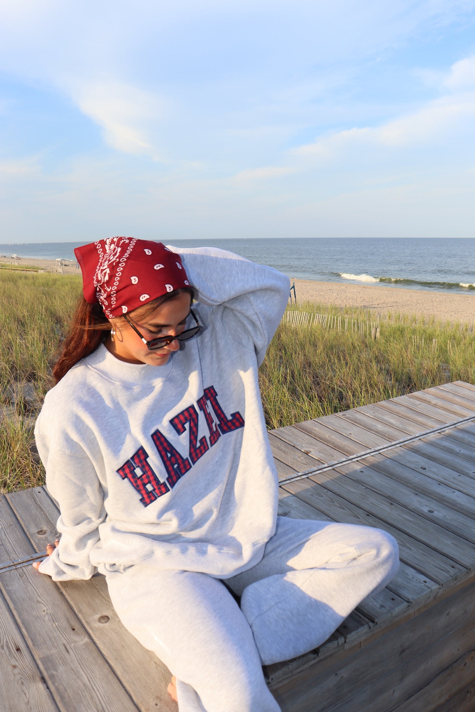 Woman in a gray HZL sweatshirt and matching pants seated on a beach boardwalk beside the ocean