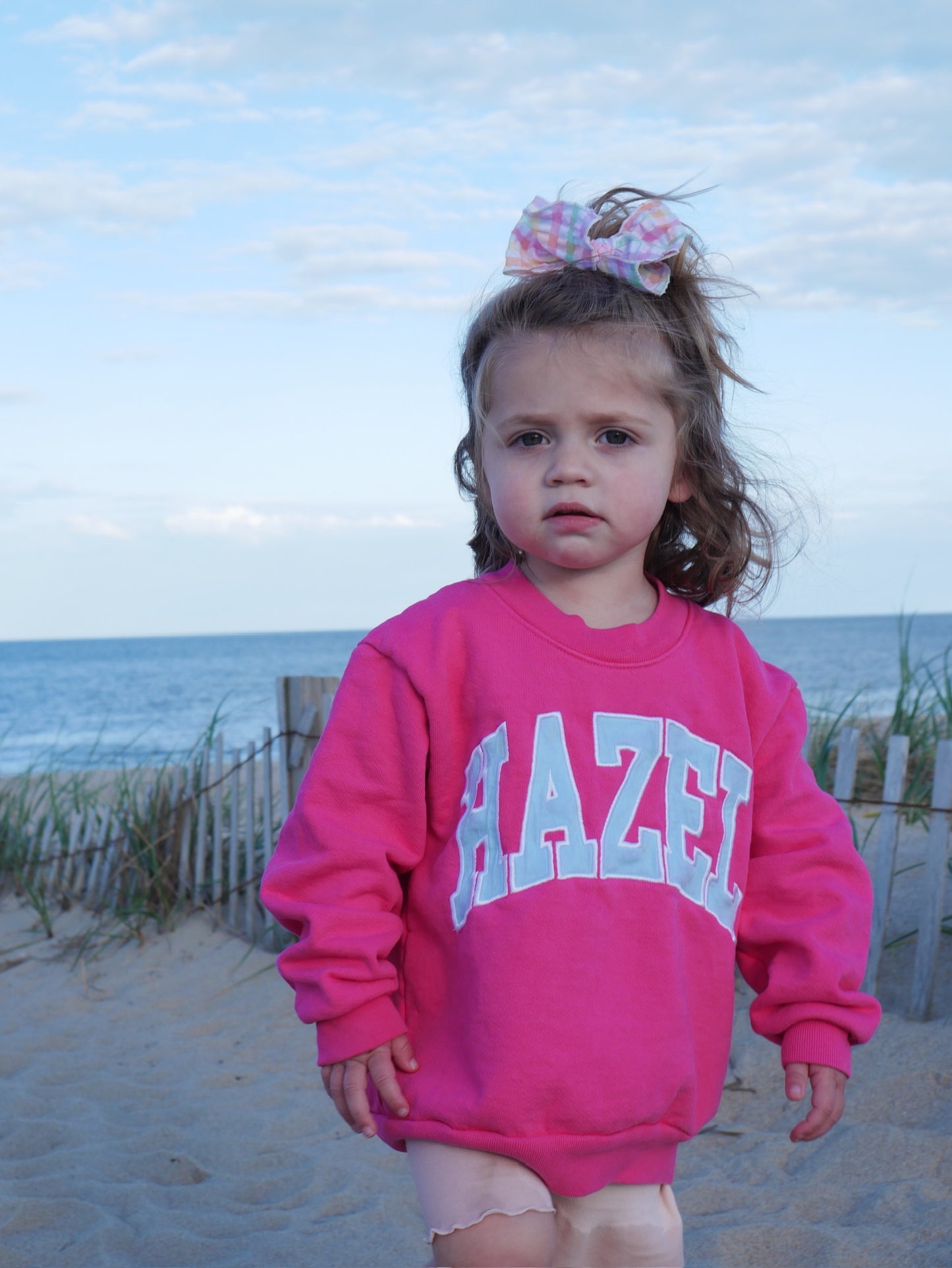 Young girl wearing a bright pink crewneck sweatshirt with white lettering standing on a sandy beach.