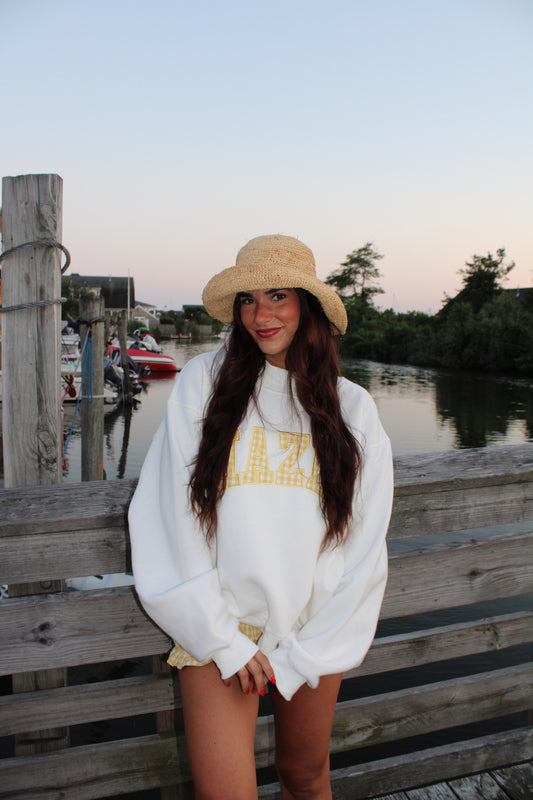 Young woman with long brown hair wearing a white oversized sweatshirt and a woven straw hat, standing on a wooden dock by the water at sunset.