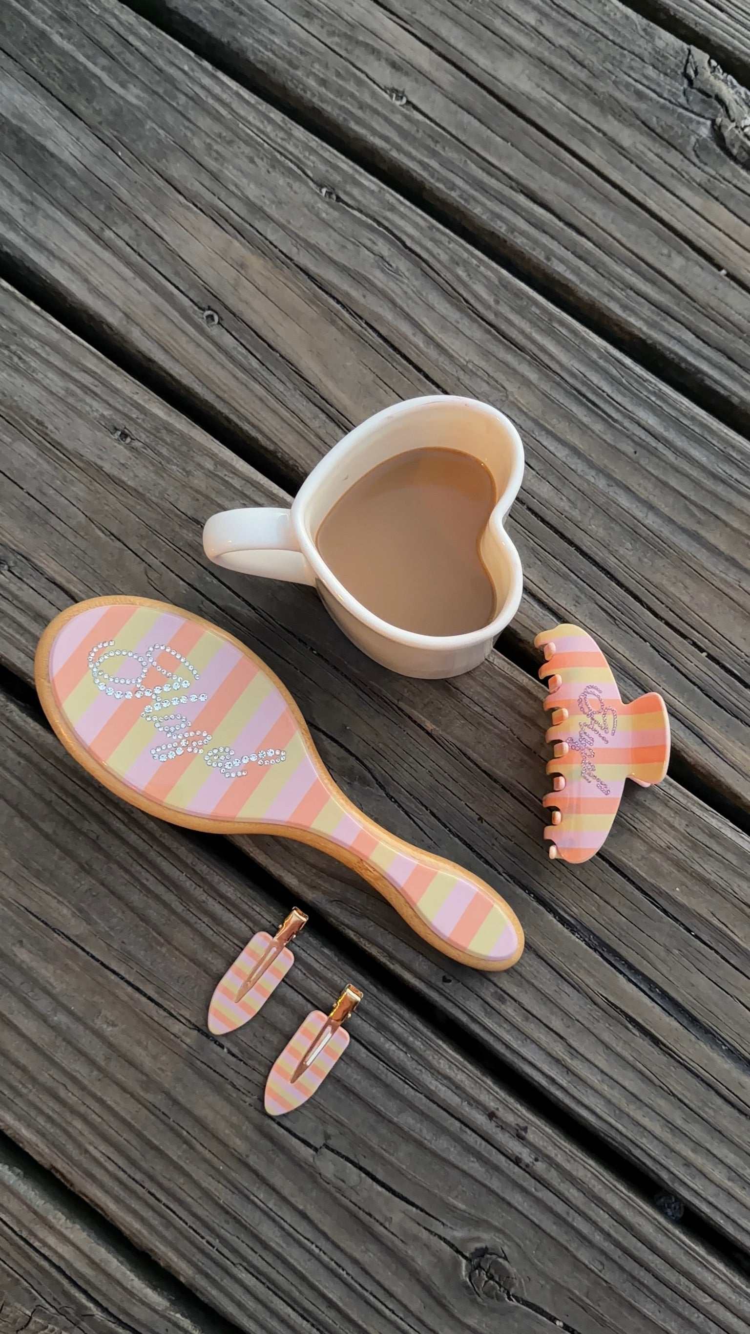 Striped pastel pink and peach hairbrush, clip, and earrings set with rhinestone details next to a heart-shaped cup of coffee on weathered wood.