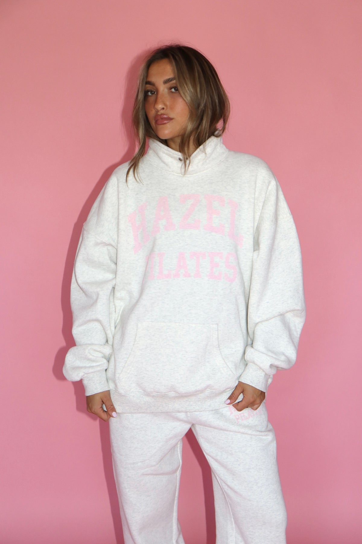 Model wearing a white Pilates sweatshirt and matching sweatpants with pink lettering against a pink backdrop