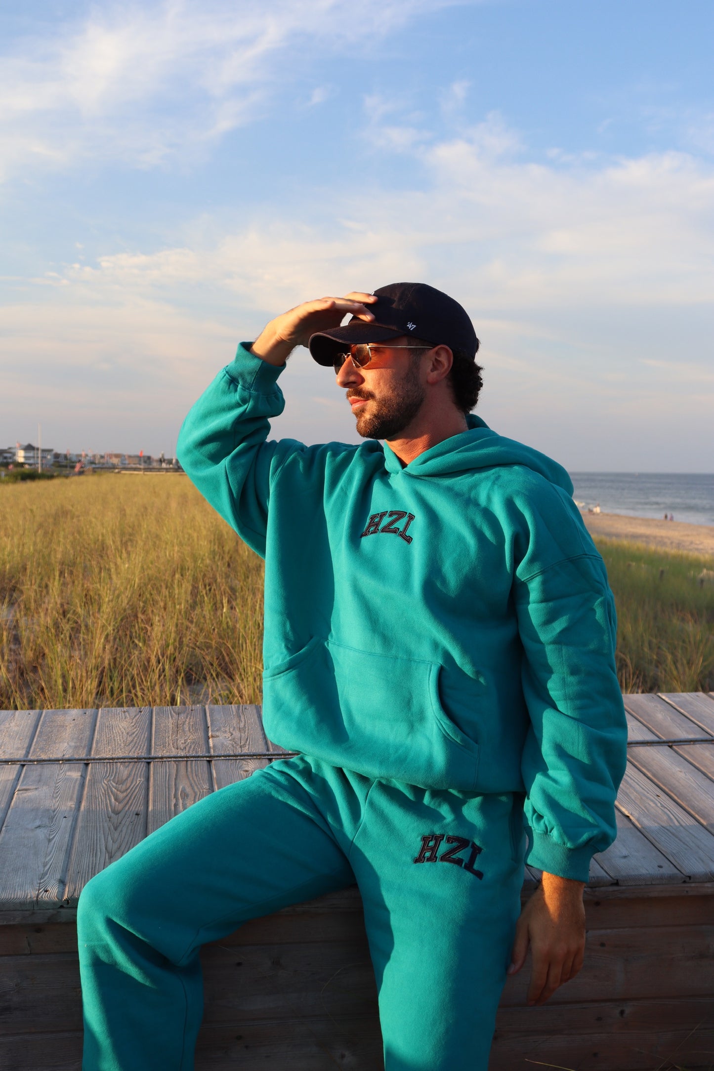 Man wearing teal HZL joggers and matching hoodie seated on a beach boardwalk at sunset