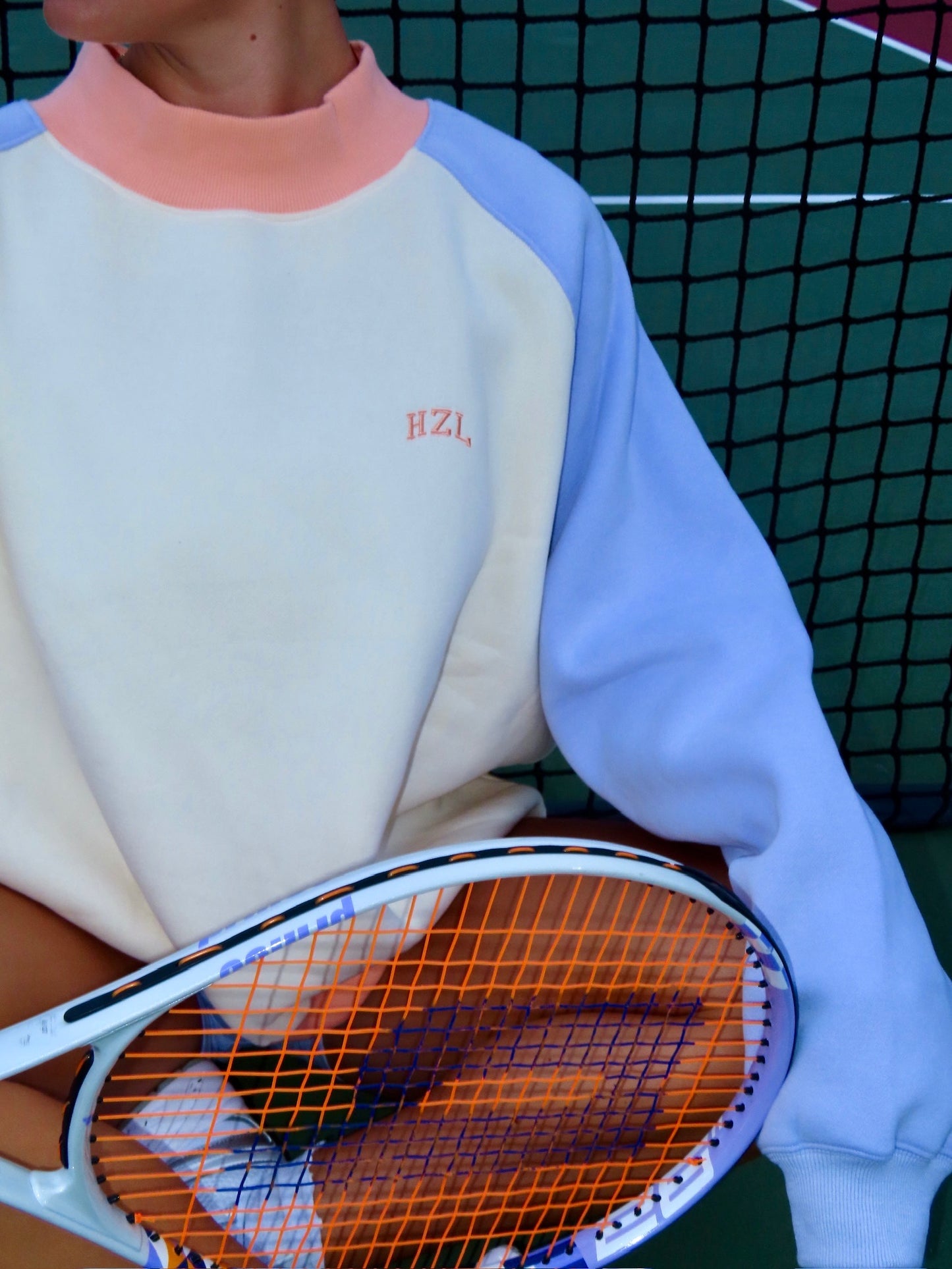 Close-up of a person wearing a pastel color-block mockneck sweatshirt holding a tennis racket in front of a net.