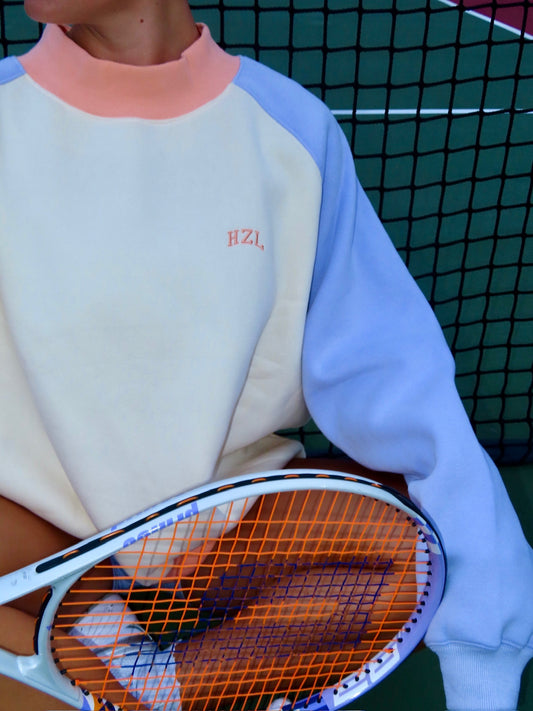 Close-up of a person wearing a pastel color-block mockneck sweatshirt holding a tennis racket in front of a net.