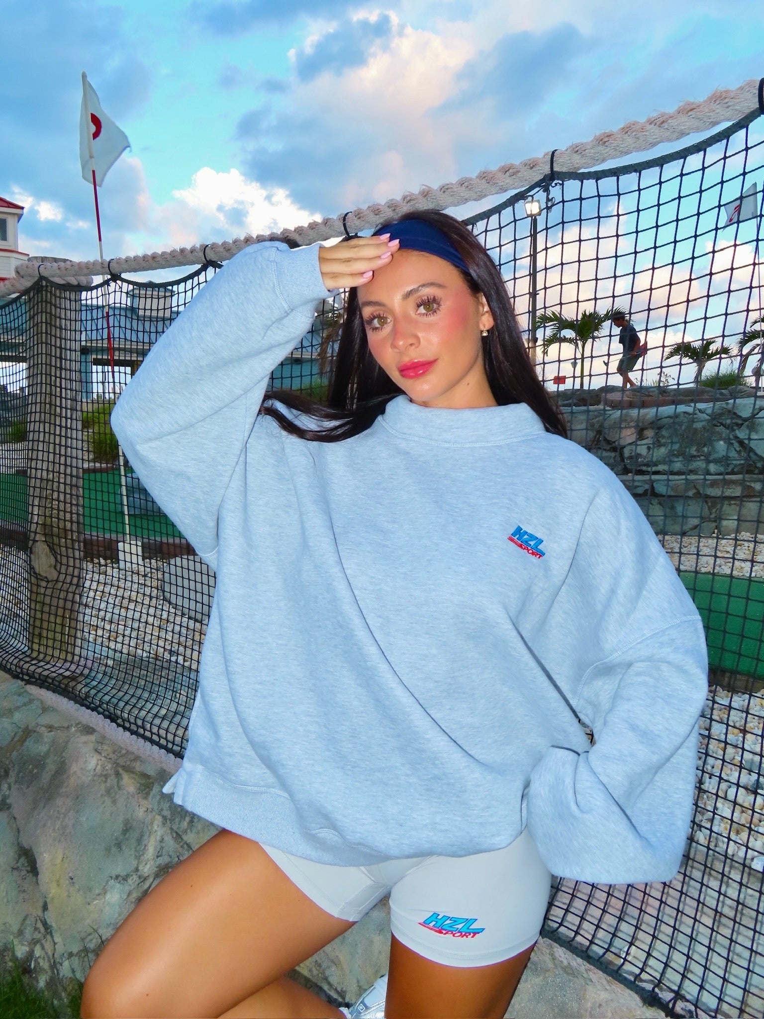 Young woman wearing a grey sporty mockneck sweatshirt and matching shorts posing near a tennis net outdoors