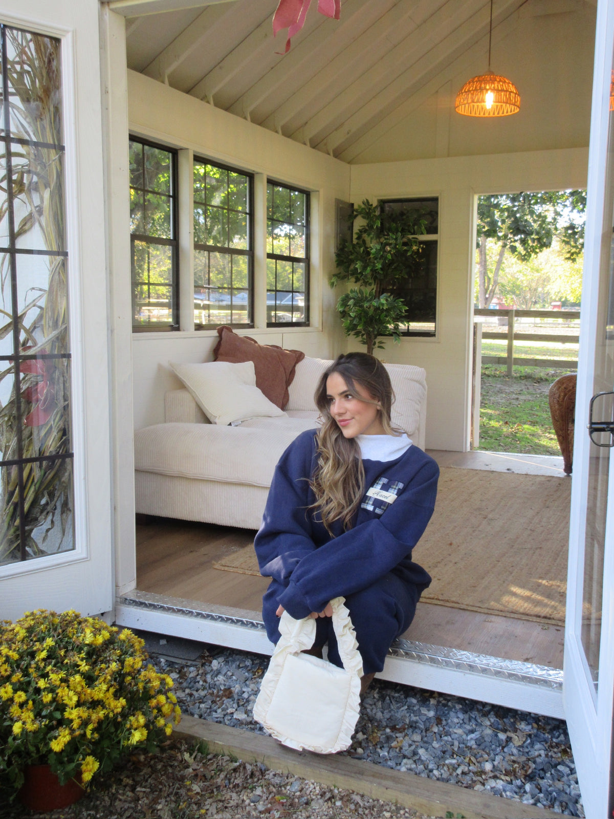 Woman wearing a navy mockneck sweatshirt seated in a cottage doorway with a white tote bag