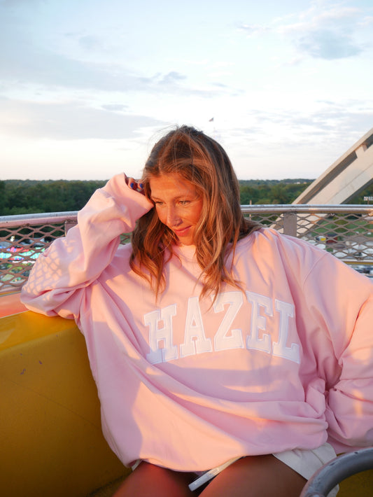 Woman wearing a soft strawberry pink oversized sweatshirt with "HAZEL" printed across the front, sitting outdoors at sunset.
