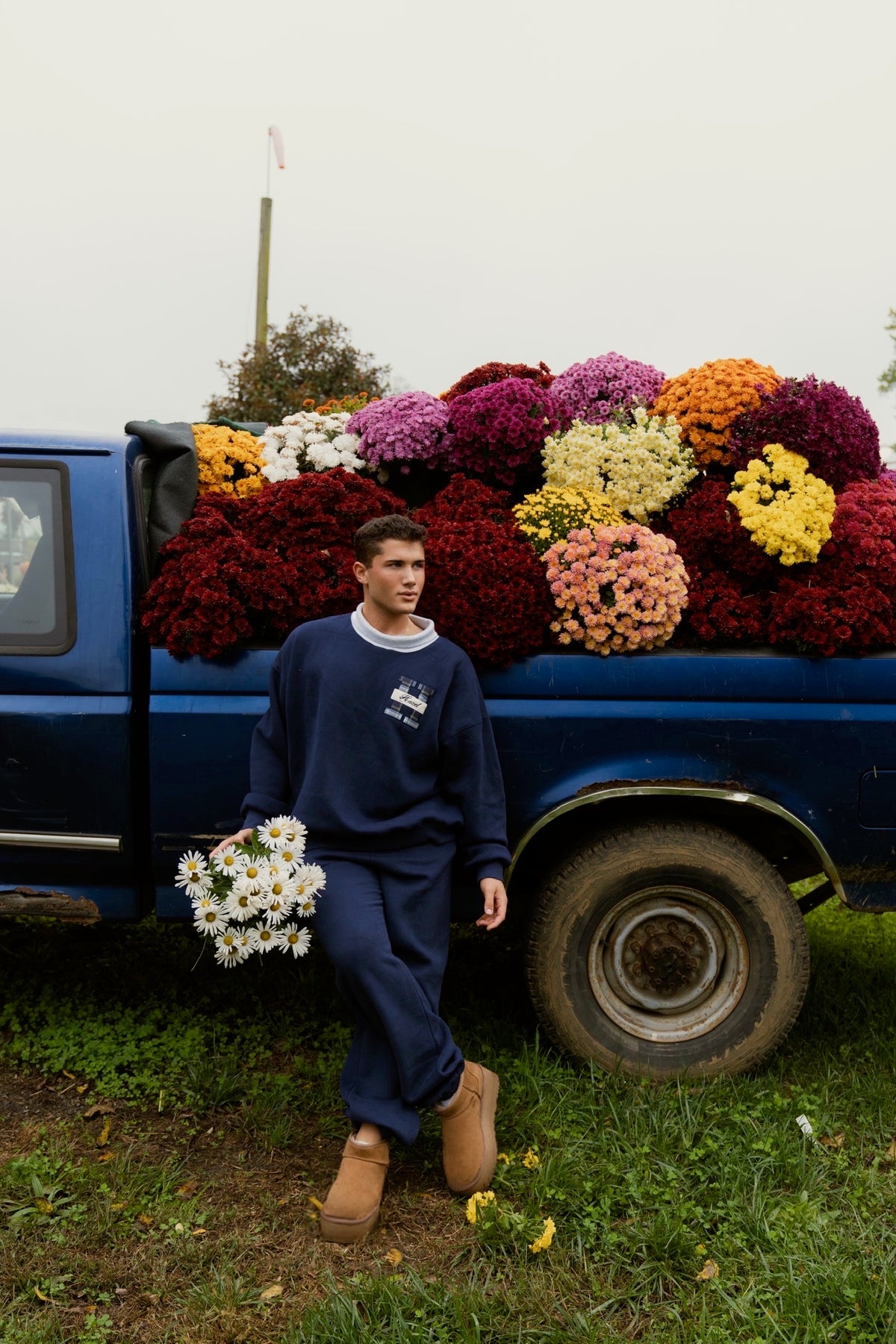 Man wearing navy joggers and a sweatshirt holding flowers beside a blue truck loaded with colorful bouquets