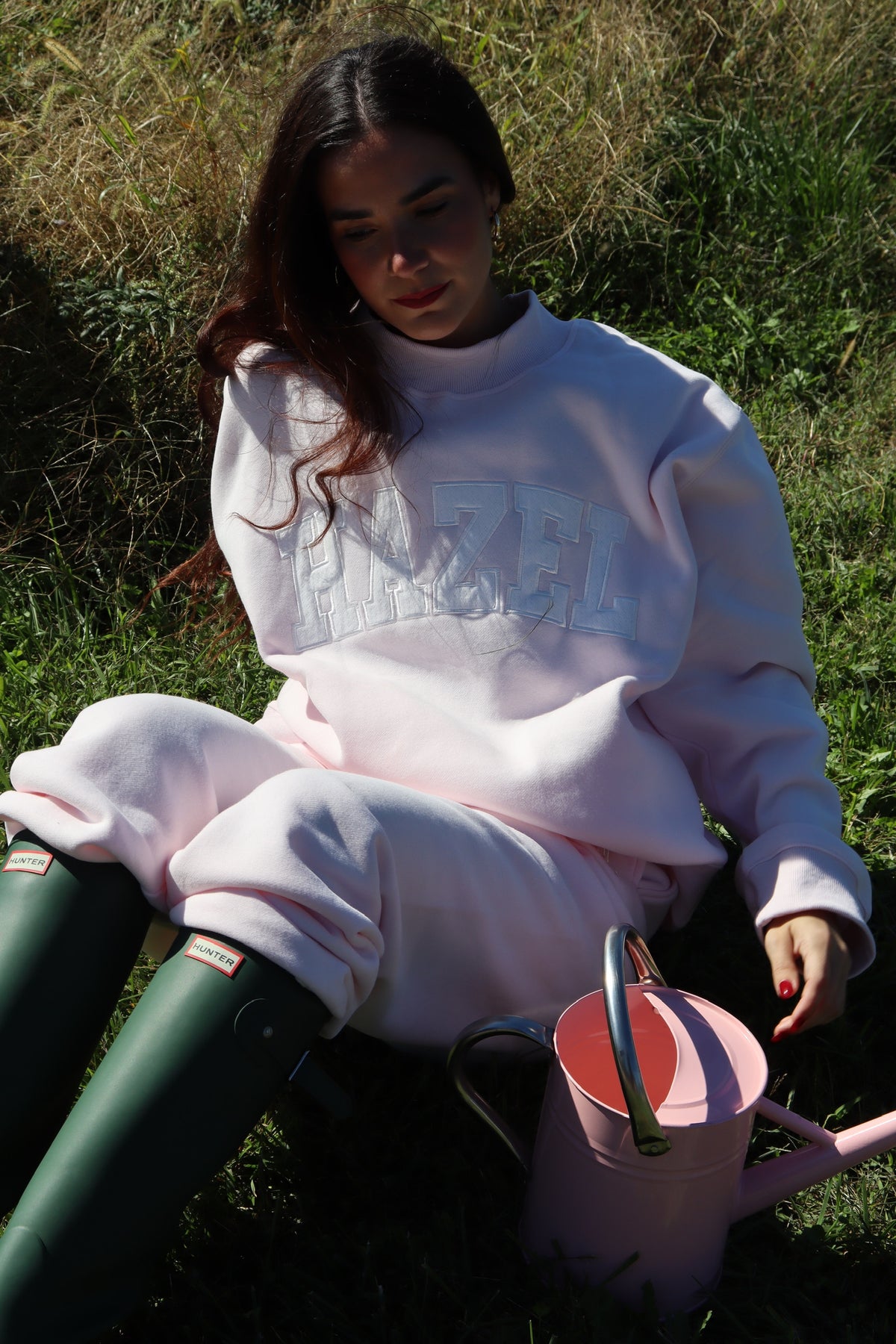 Woman wearing a pale pink mockneck sweatshirt and matching pants beside a pink watering can outdoors