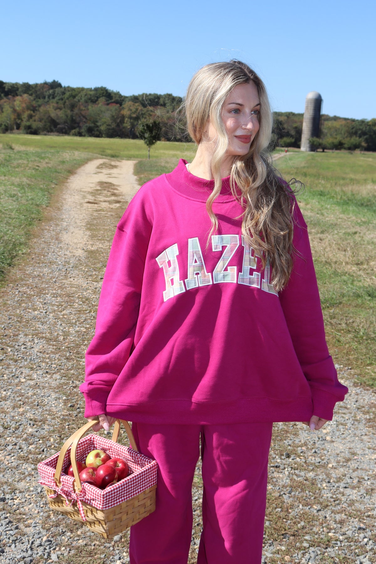Woman wearing a hot pink mockneck sweatshirt and matching pants while carrying an apple basket outdoors