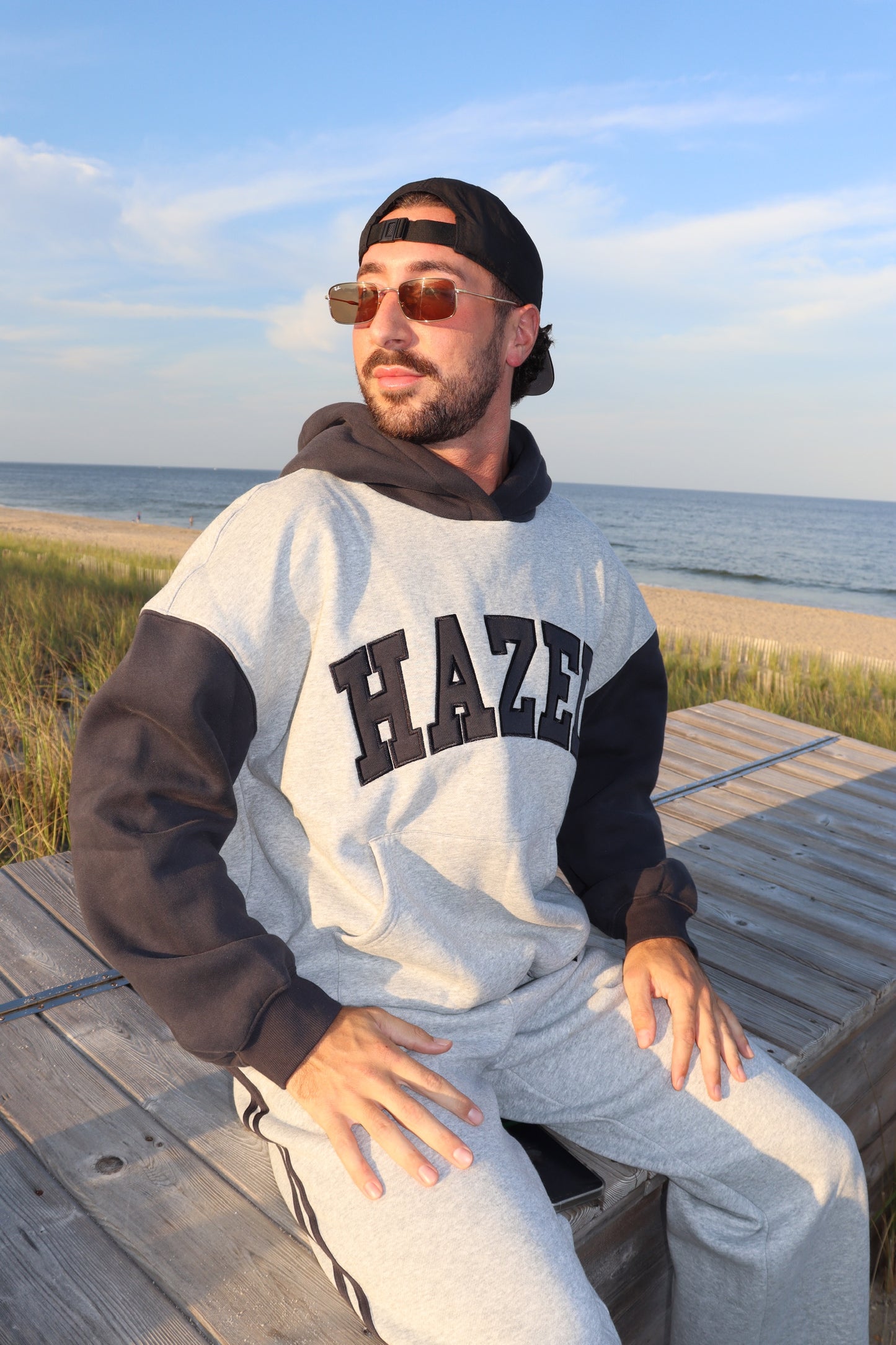 Man wearing a gray HAZEL graphic hoodie and sunglasses seated on a beach boardwalk