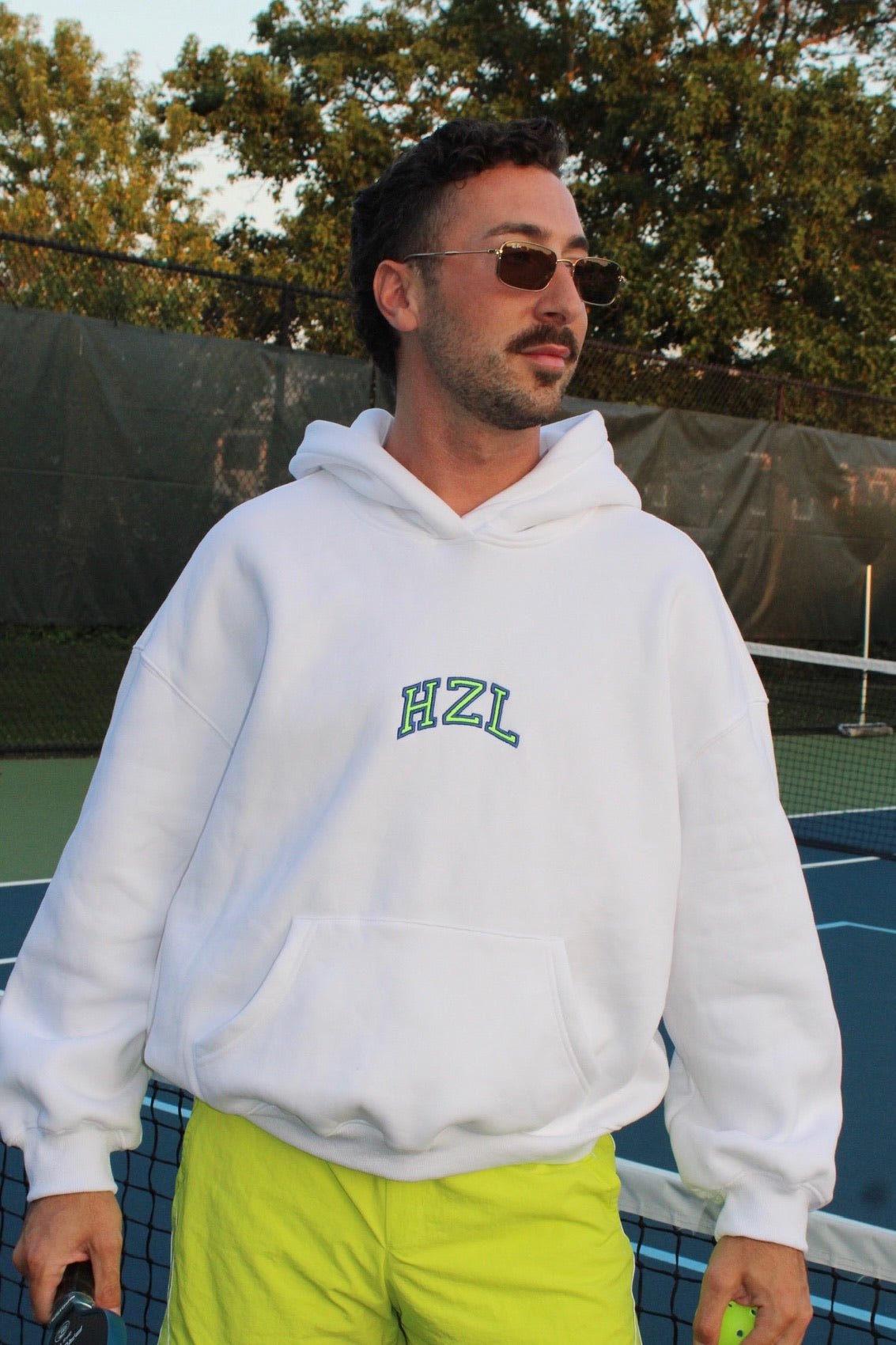 Man wearing a white hoodie with "HZL" logo and bright yellow shorts standing on a tennis court holding a ball.