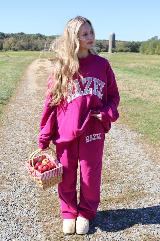 Model wearing a magenta HZL sweatshirt and matching sweatpants while carrying a basket of apples outdoors