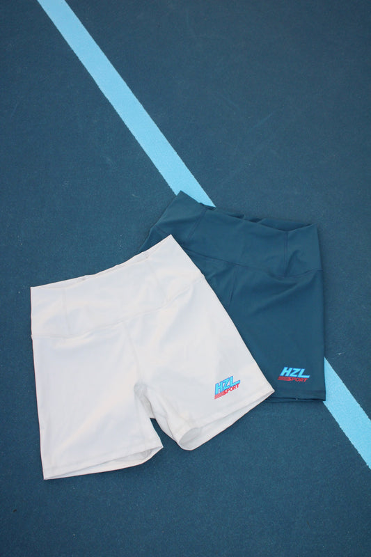 Two pairs of sporty shorts in cream and dark teal colors with a small HZL Sport logo on the leg, laid flat on a blue court surface.