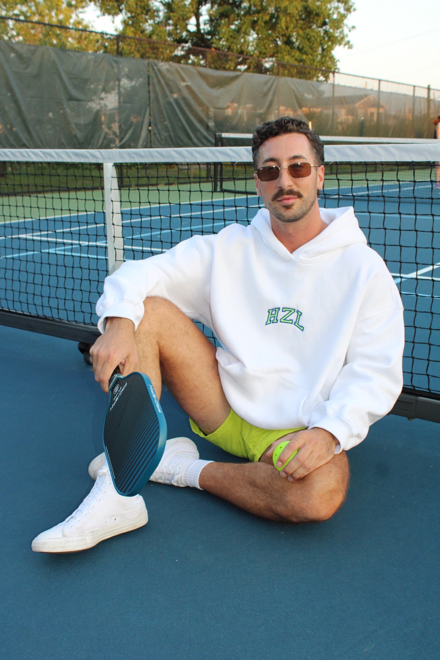 Man wearing a white hoodie with green lettering sitting on a tennis court holding a paddle and ball, casually styled with sunglasses and white sneakers.