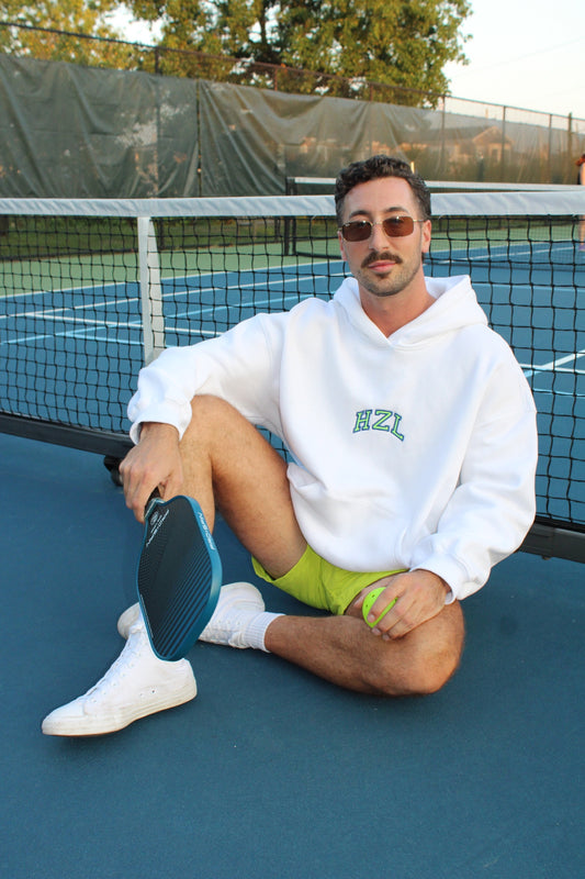 Man wearing a white hoodie with green lettering sitting on a tennis court holding a paddle and ball, casually styled with sunglasses and white sneakers.