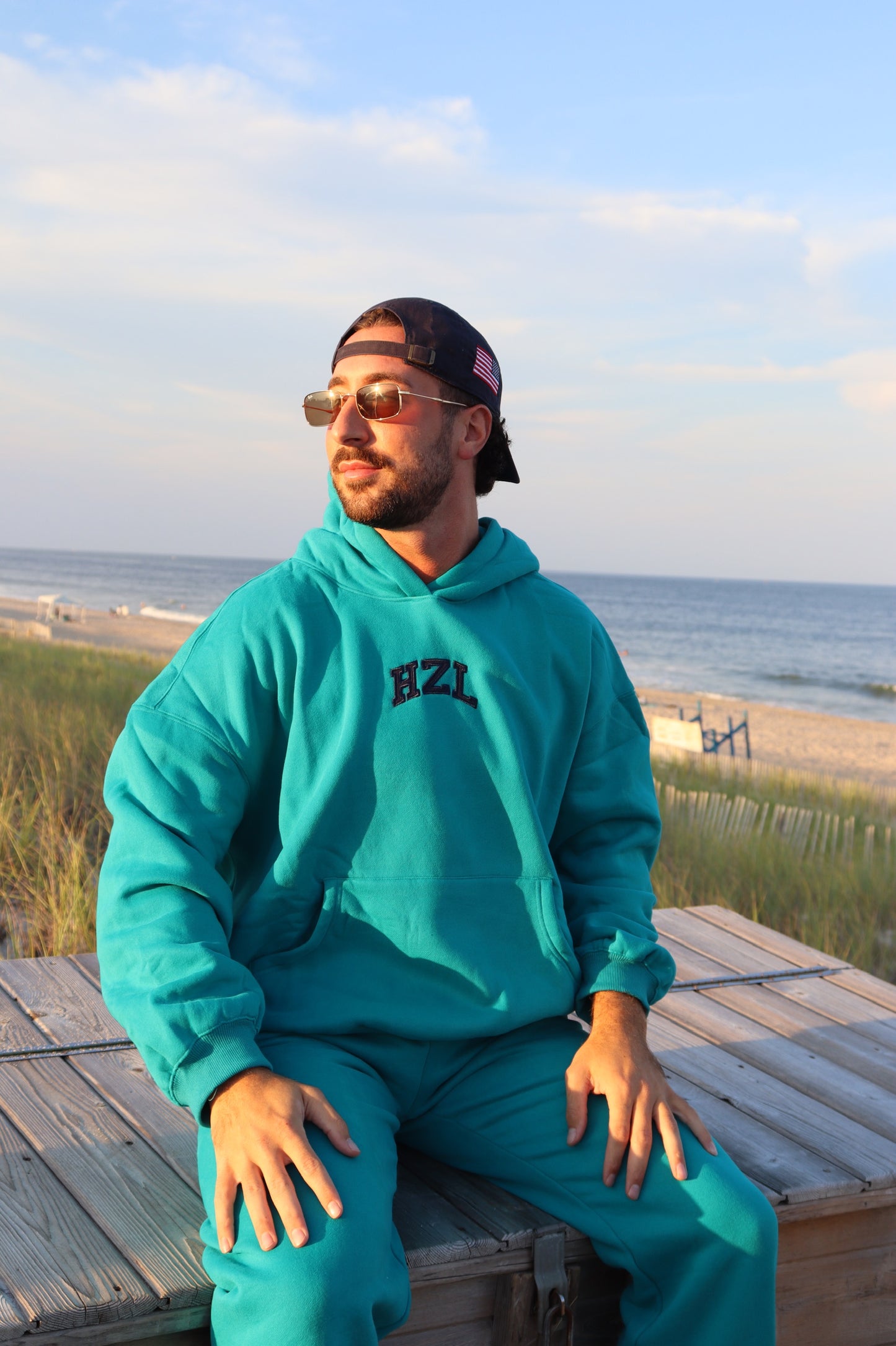 Man wearing a turquoise HZL hoodie and matching pants seated on a beach boardwalk by the ocean